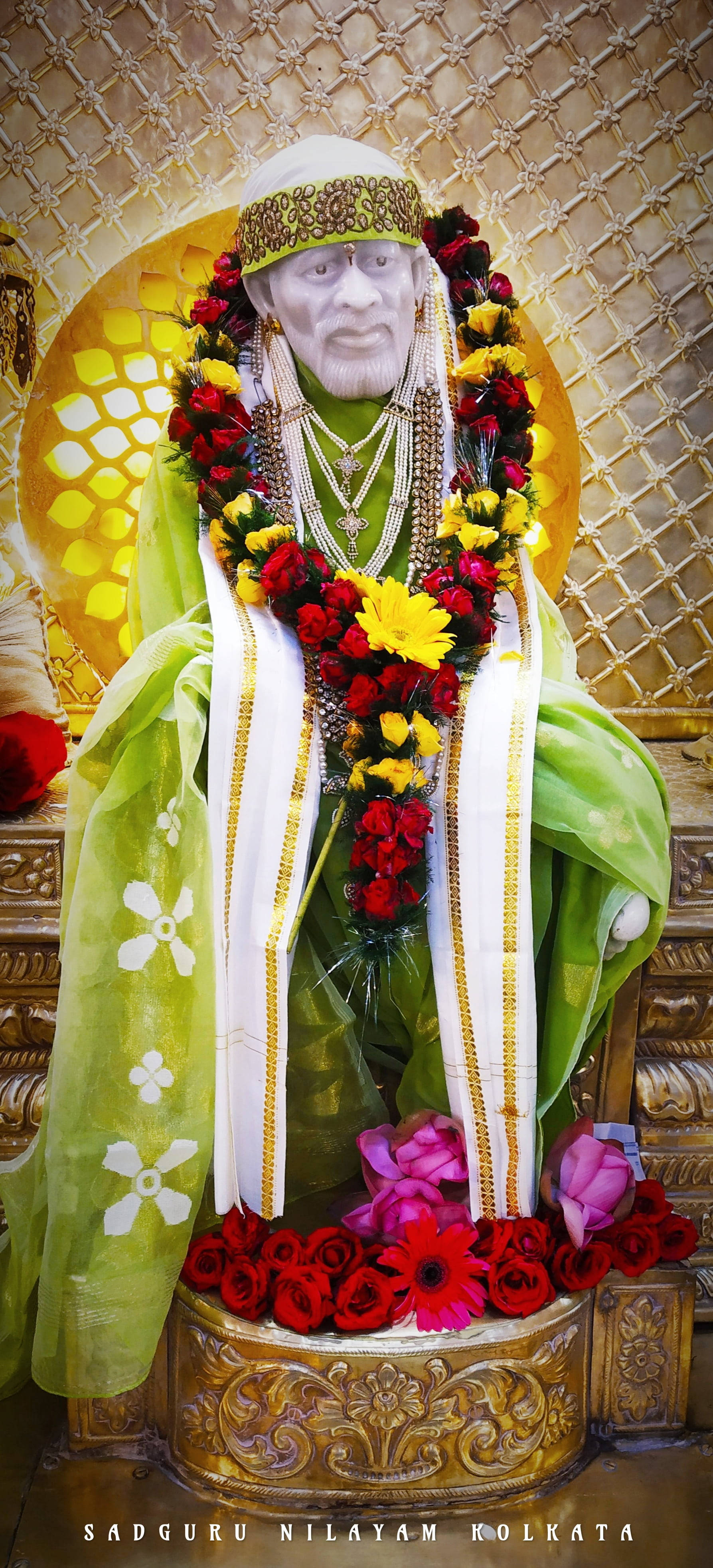 身穿綠袍的聖靈 Sai Baba 手機壁紙 壁紙 — 一座寧靜的Sadguru Nilayam Kolkata雕像，飾以鮮豔的紅色和黃色花朵，並披上綠色和白色布料，背景為金色格子。