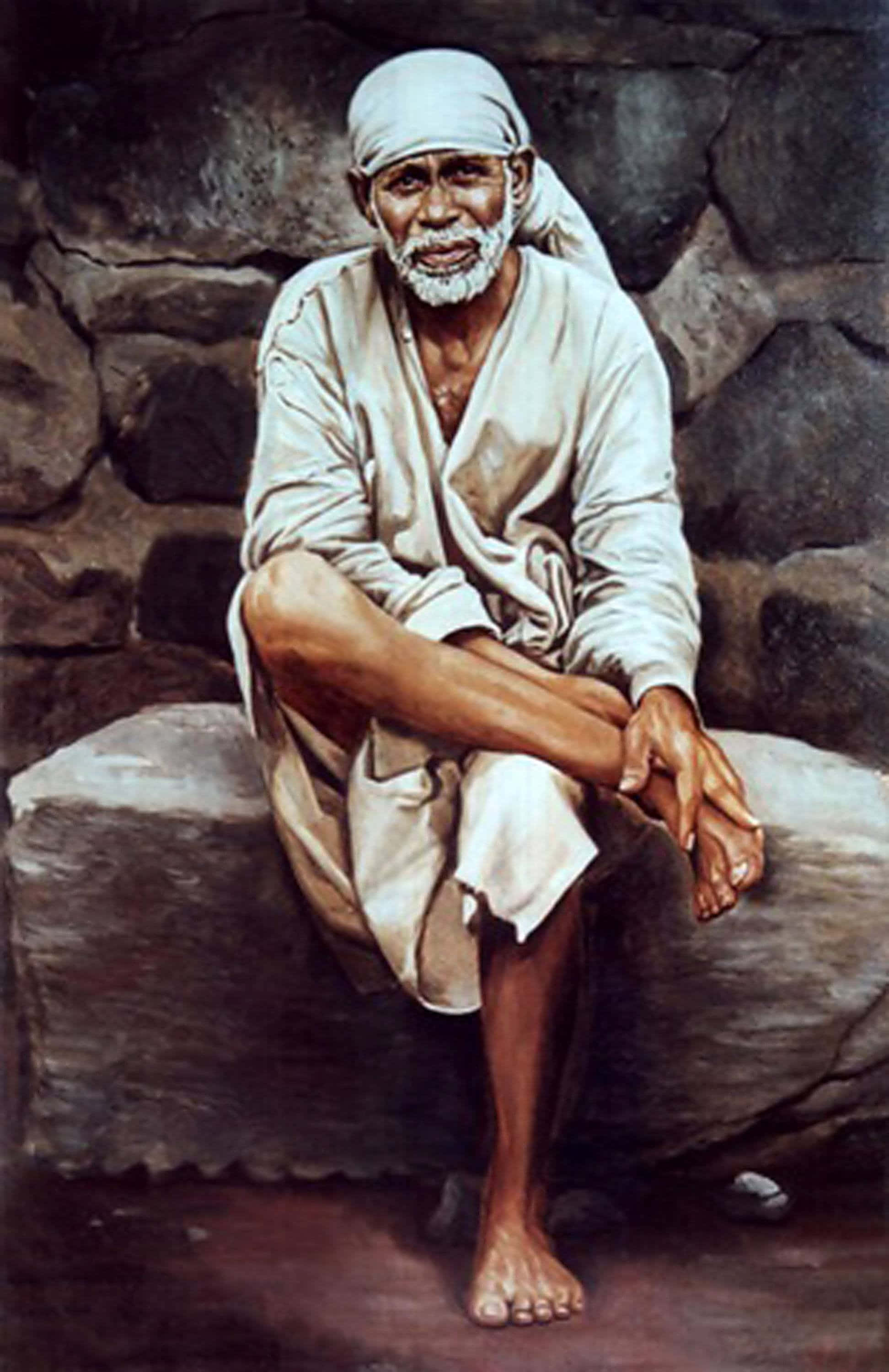 Sai Baba 坐在岩石上的畫像 壁紙 — 一位留著白鬍子、戴頭巾的長者赤腳坐在石壁前的石台上。