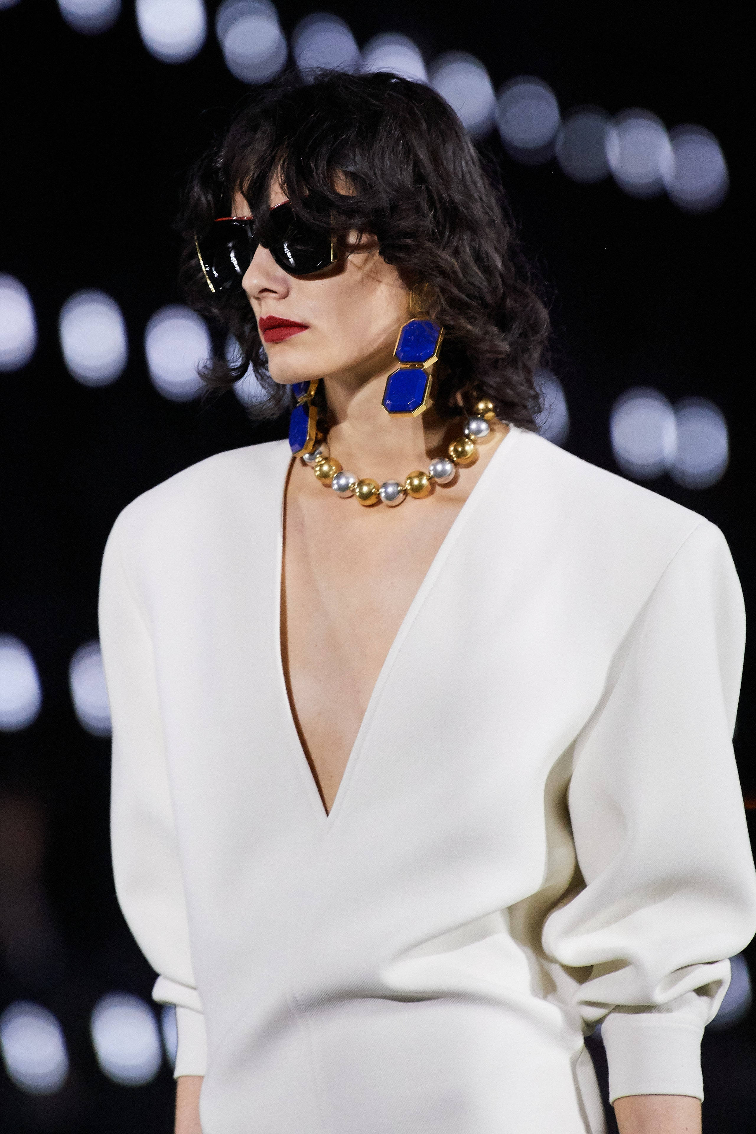 Saint Laurent 2022春夏系列 壁紙 — 一位擁有深色捲髮和大膽藍色耳環的模特兒，在閃爍的散景燈光下自信地穿著白色長裙行走。