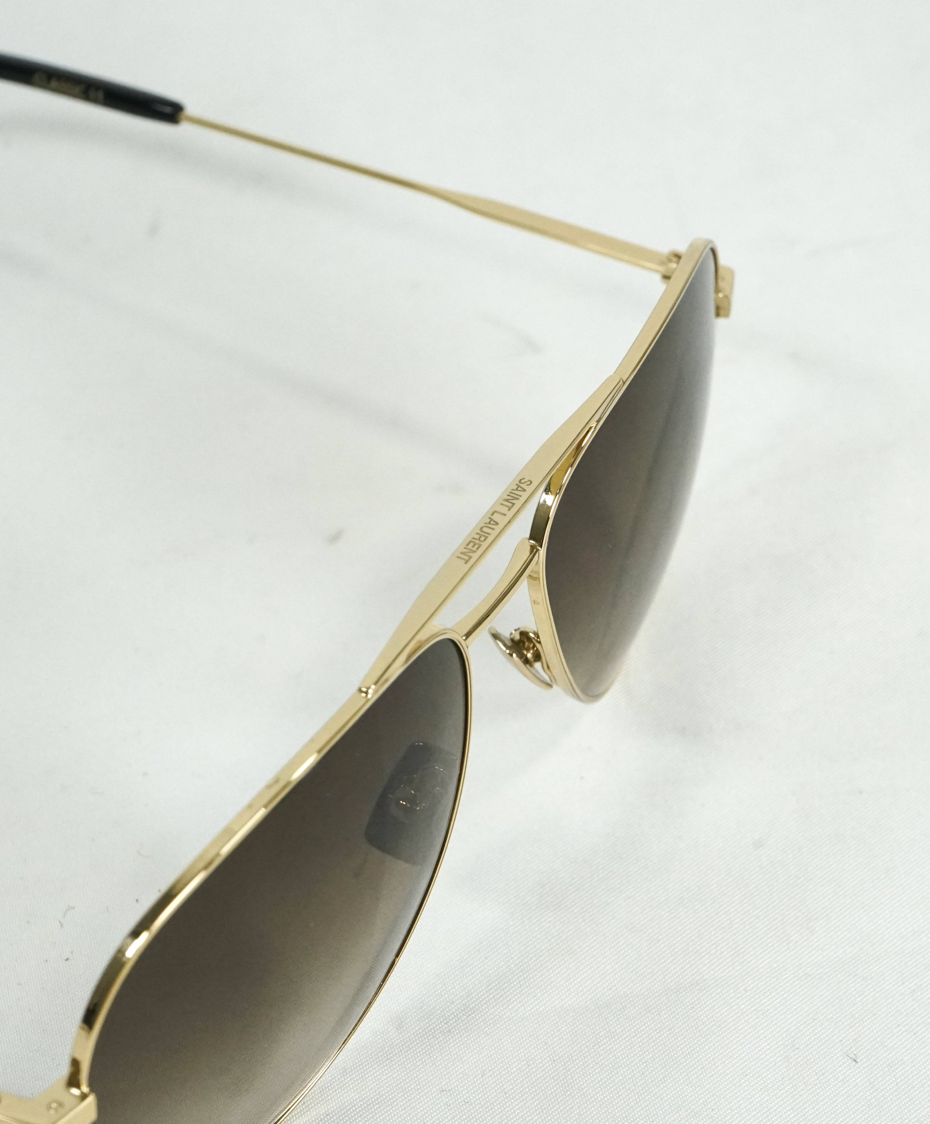 Saint Laurent 金屬金框男款飛行員太陽眼鏡 壁紙 — 金色框飛行員太陽眼鏡特寫，深色鏡片，放在白色紋理表面上。