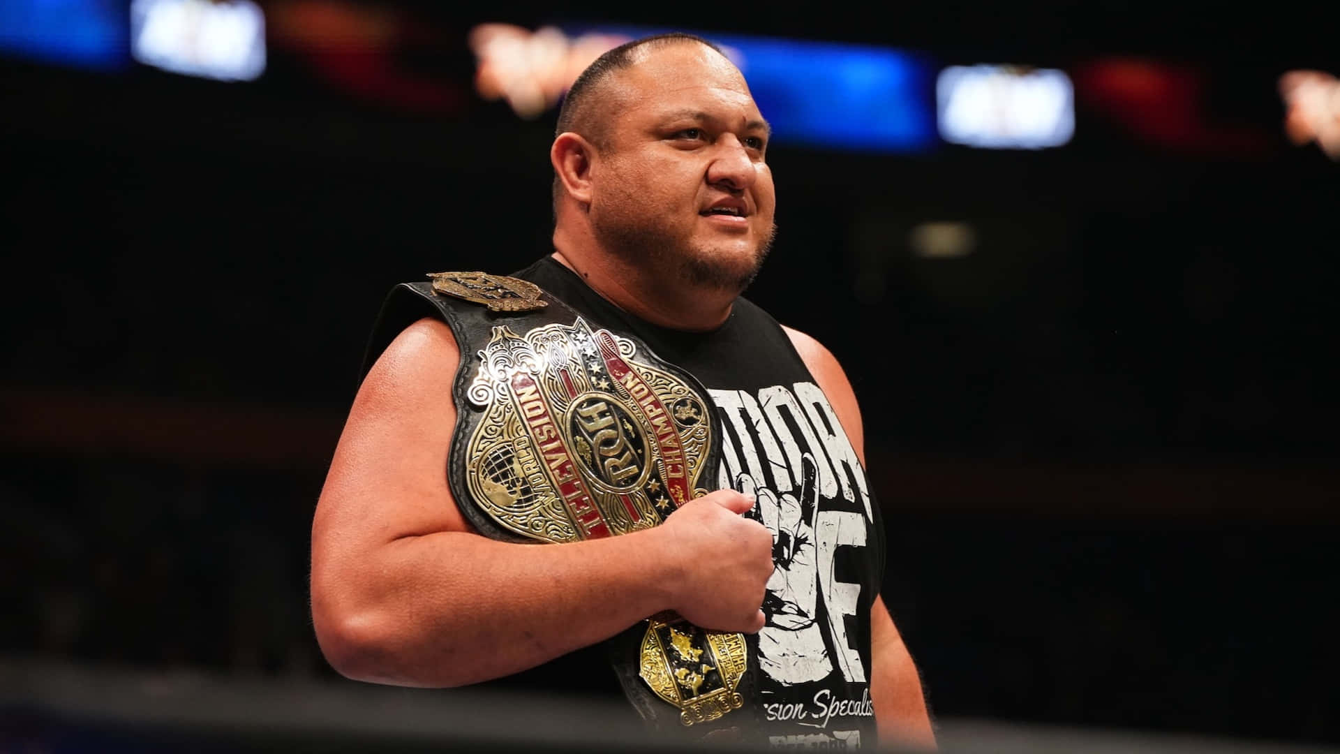 Samoa Joe AEW 冠軍腰帶肩掛壁紙 壁紙 — 一位肌肉發達的摔角手穿著黑色背心，在場館燈光下自豪地舉起閃亮的冠軍腰帶。