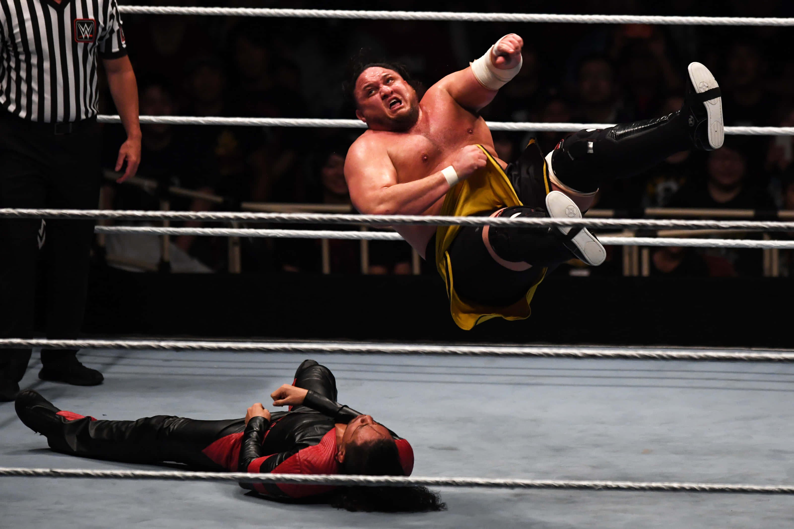 Samoa Joe 對決 Shinsuke Nakamura WWE 2019 東京現場 壁紙 — 一名穿著黃色短褲的摔角手在空中躍向摔角場內躺在墊子上的對手。