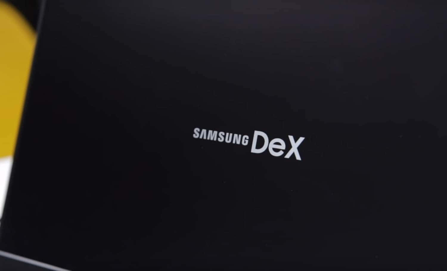 Samsung DeX 標誌 壁紙 — Samsung DeX 標誌的特寫，白色文字印在光滑的黑色表面上，帶有黃色背景元素。