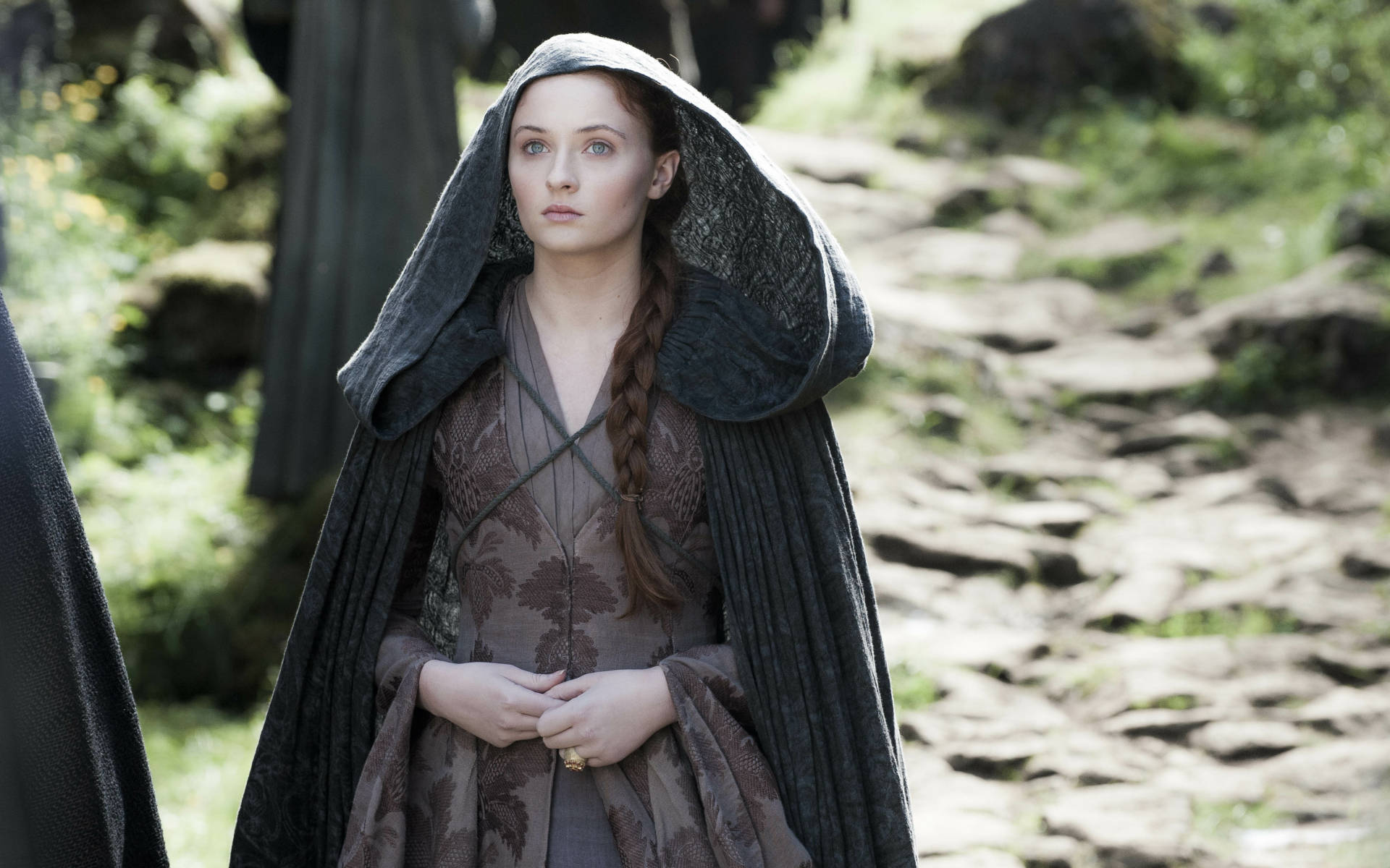 Sansa Stark《冰與火之歌》第四季 壁紙 — 一位身穿深色連帽斗篷的女子站在陽光普照的石徑上，穿越一片鬱鬱蔥蔥的綠色森林。