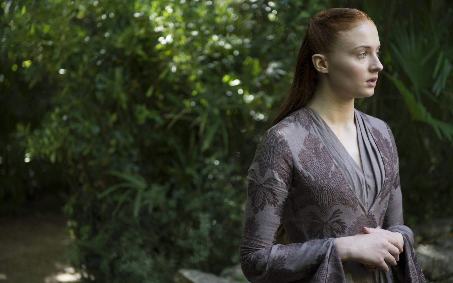 Sansa Stark穿紫色禮服 壁紙 — 一位紅髮女子身穿灰色圖案長袍，站在翠綠的花園中，若有所思地望向一旁。