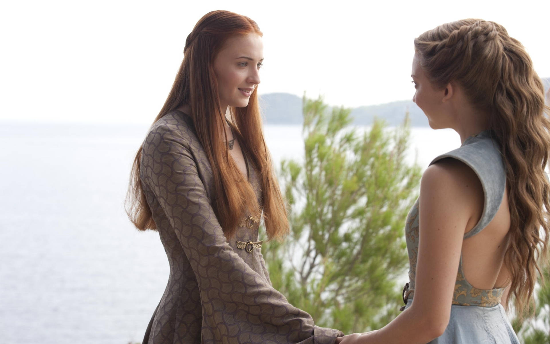 Sansa Stark與Margaery Tyrell 壁紙 — 兩位身穿中世紀風格連衣裙的女性手牽手，在綠意盎然和水邊的海岸背景下相視而笑。