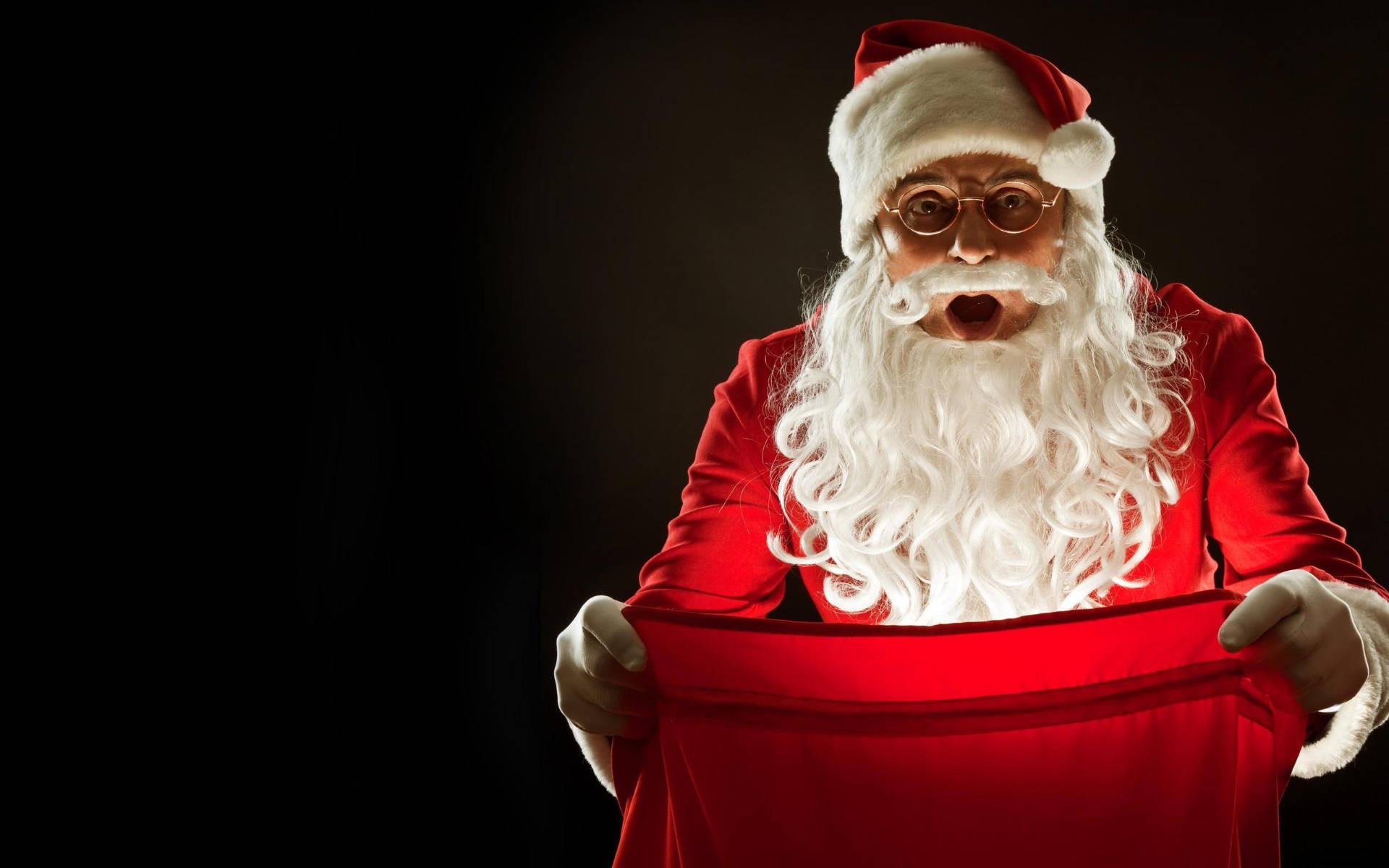 Santa Claus With Gift Bag 壁紙 — 戴眼鏡的聖誕老人帶著驚訝的表情，在深色背景前打開一個紅色袋子。