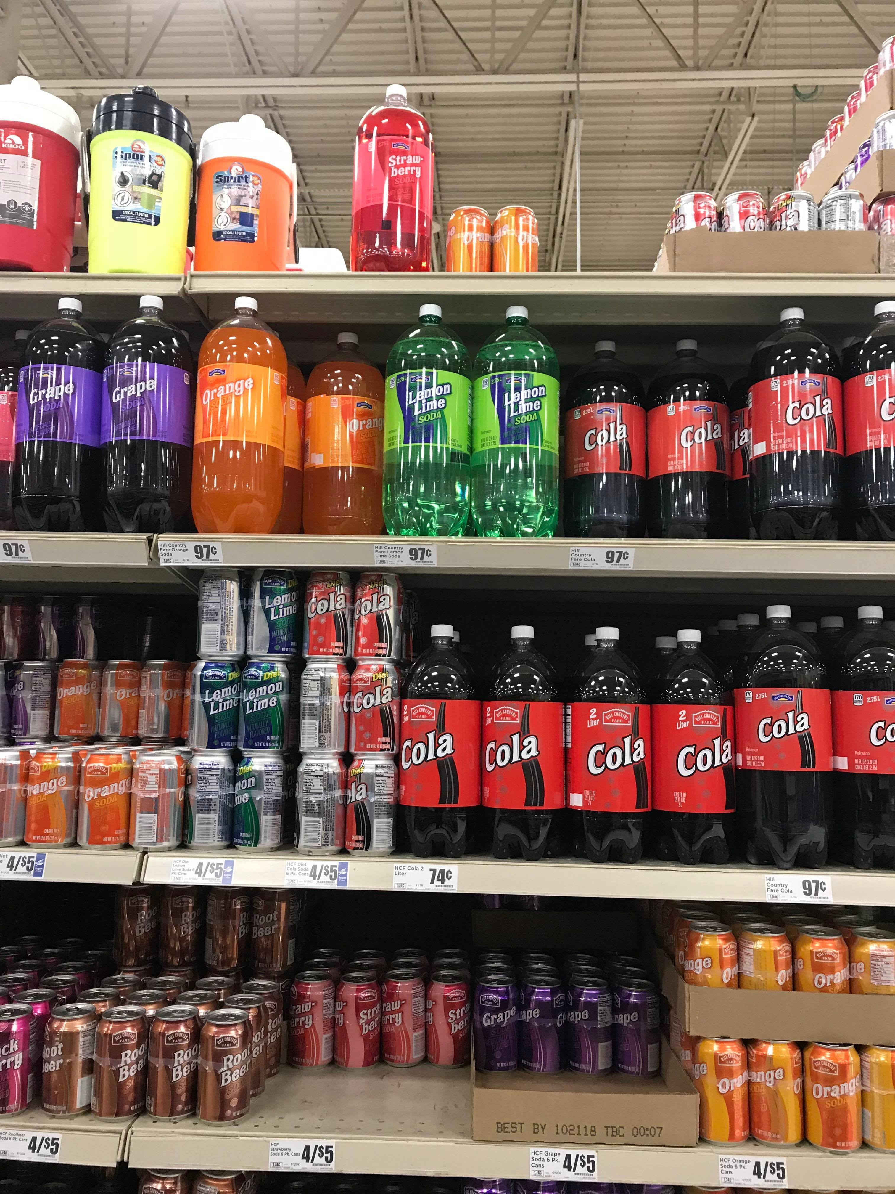 Save A Lot 超市碳酸飲料 壁紙 — 一個貨品齊全的超市貨架展示著一排排色彩繽紛的汽水瓶和罐頭，在螢光燈下呈現鮮豔色調。