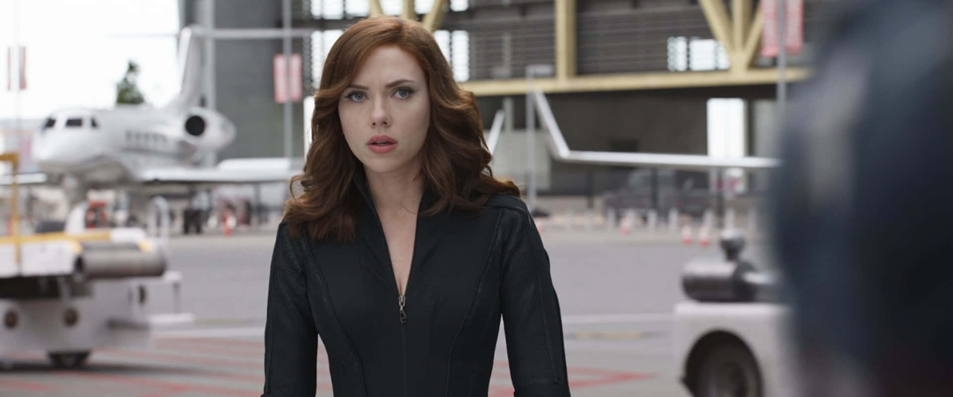 黑寡婦 桌布 壁紙 — Scarlet Witch 站在機場停機坪，身穿黑色外套，背景有私人飛機和地面車輛。