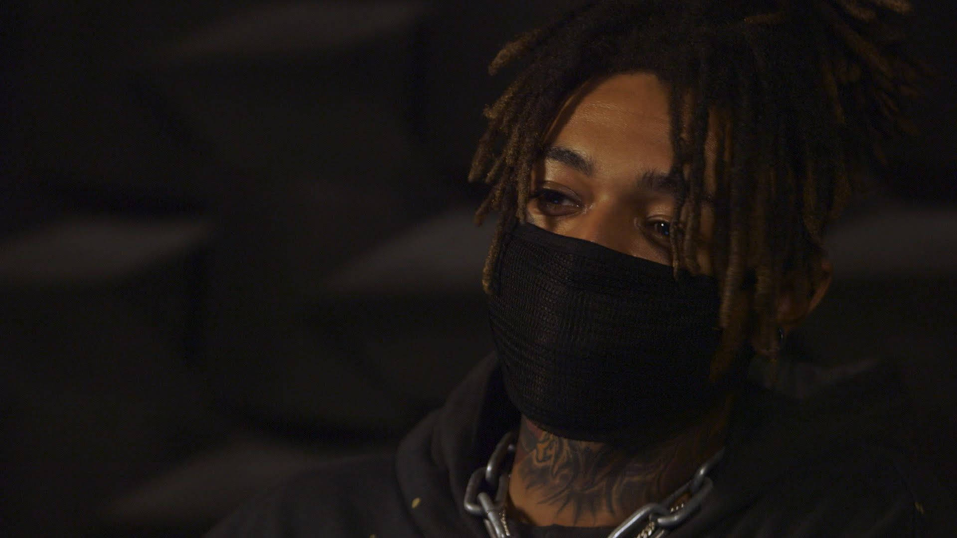 Scarlxrd 戴口罩 壁紙 — 一個留著辮子頭、戴黑色口罩的人，在昏暗燈光下若有所思地凝視，可見紋身和鏈條項鍊。