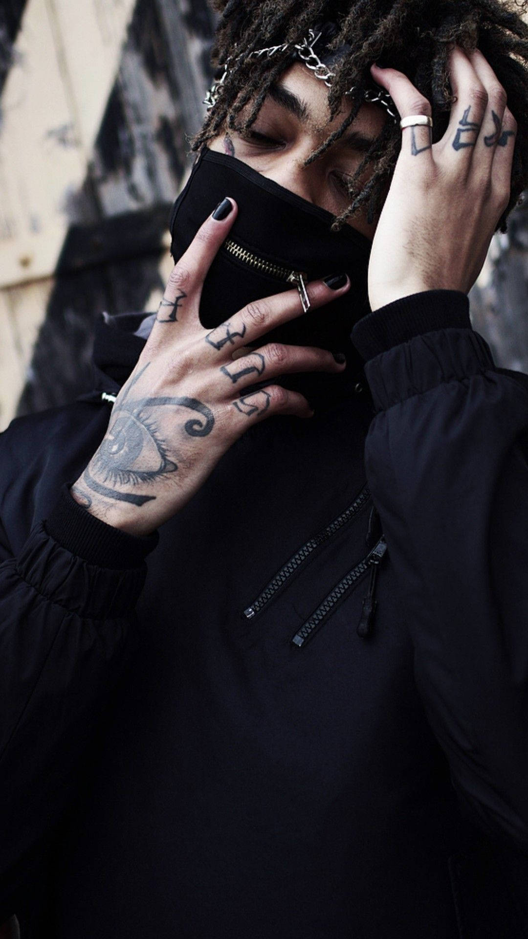 Scarlxrd 手部紋身 壁紙 — 一個有辮子頭和紋身的人正在調整黑色口罩，背景是粗糙的城市環境。