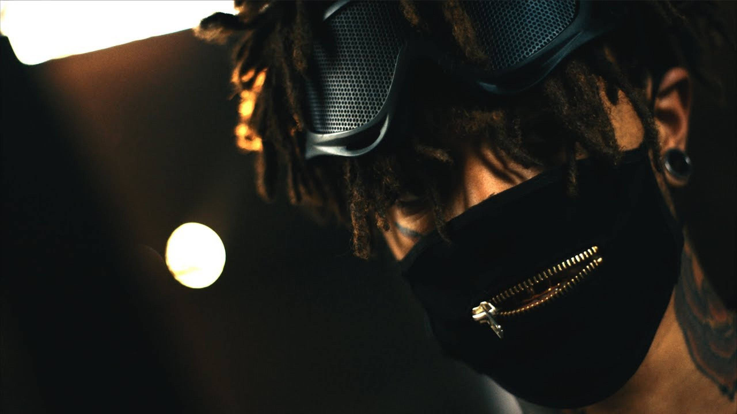 Scarlxrd 拉鍊口罩 壁紙 — 一個留著辮子的人的特寫，戴著黑色拉鍊面罩和網眼護目鏡，在溫暖的電影燈光下。