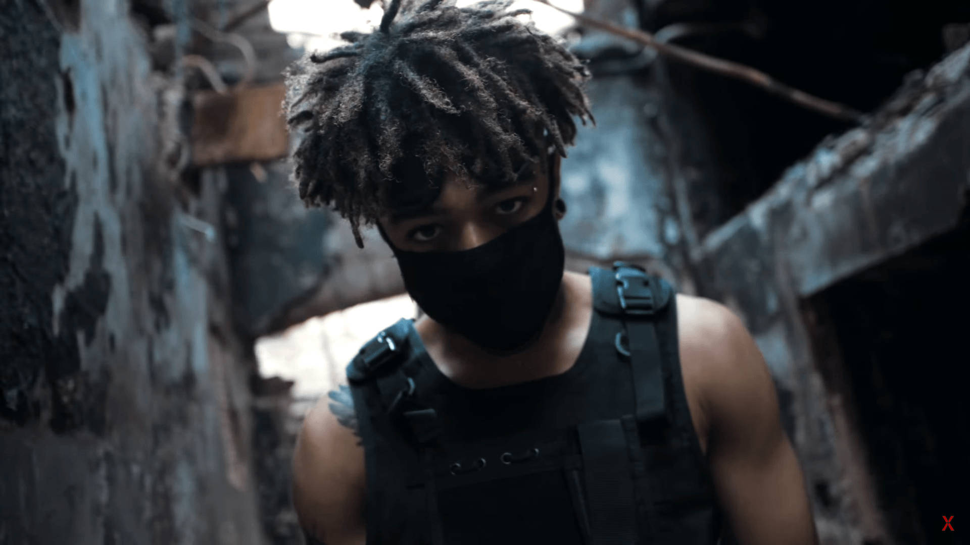 Scarlxrd 搭配戰術背心 壁紙 — 一個戴面具、梳著辮子的人在昏暗燈光下從破爛的小巷中凝視著。