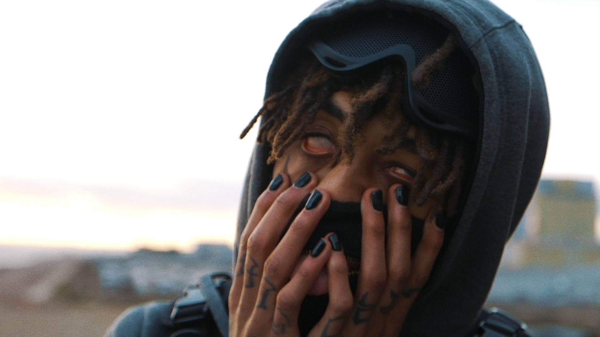 Scarlxrd 眼球上翻 壁紙 — 一個留著辮子、塗黑色指甲油的人，用紋身的手遮住嘴巴，穿著連帽衫和護目鏡，背景是朦朧的城市景觀。
