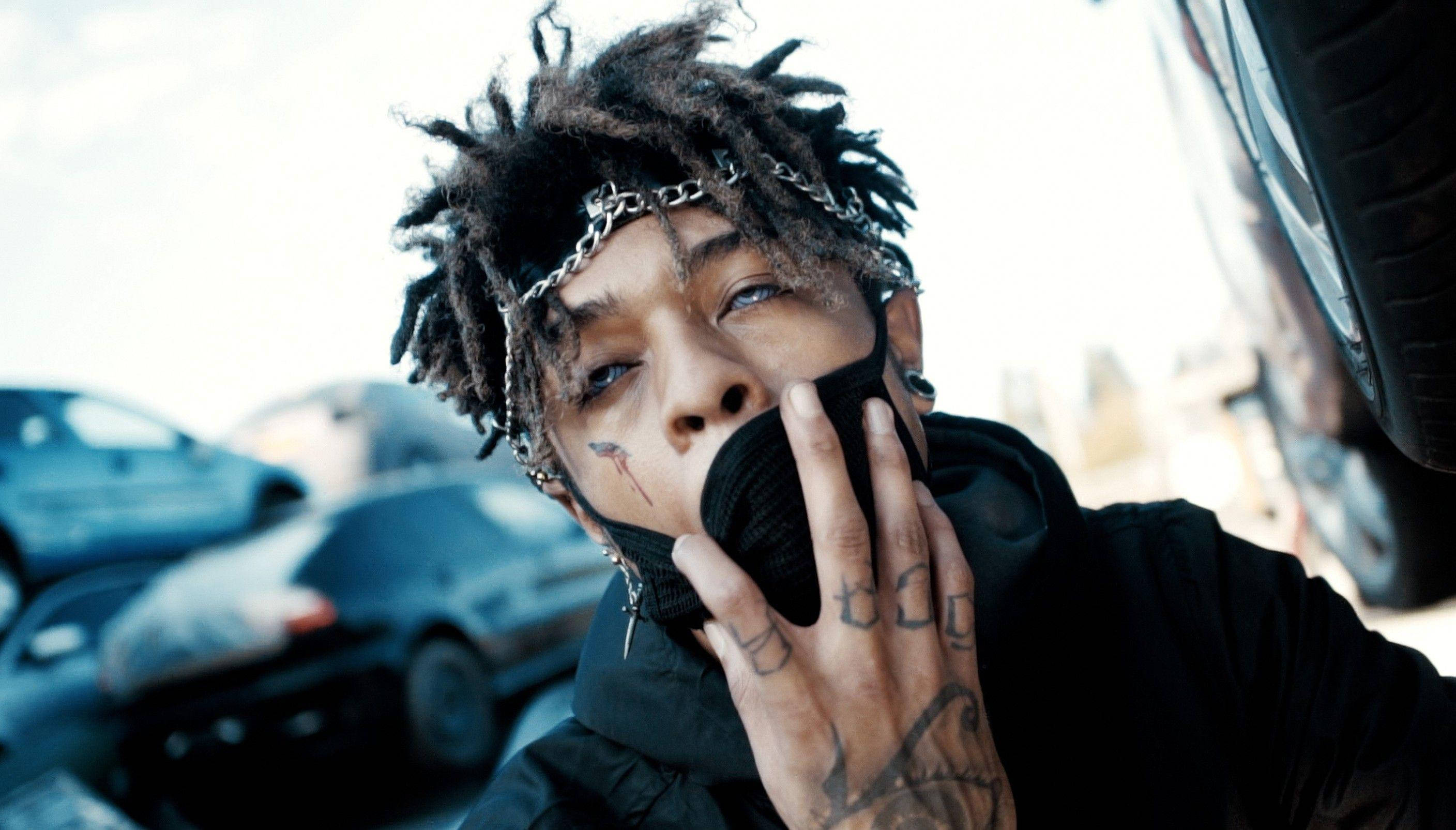 Scarlxrd 着寫輪眼鏡 壁紙 — 一名年輕男子留著辮子頭和紋身，在粗糙的城市環境中站在汽車附近，用黑色手套捂住嘴巴。