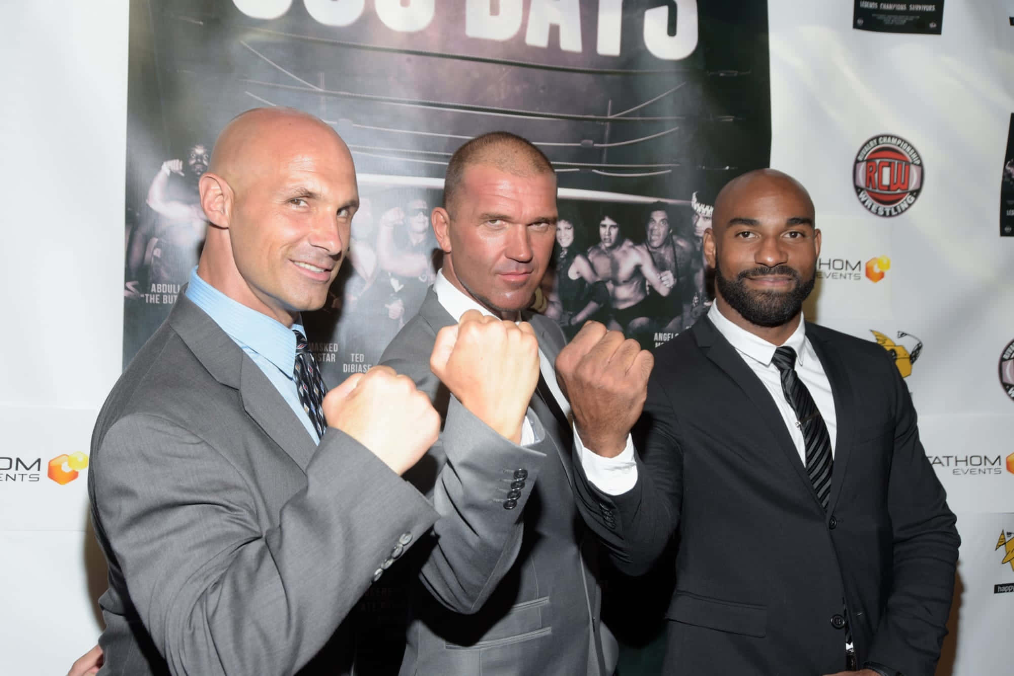 Scorpio Sky、Frankie Kazarian 與 Christopher Daniels 身著禮服合影 壁紙 — 三位光頭男士身穿西裝，在拳擊主題背景前舉起拳頭擺姿勢。