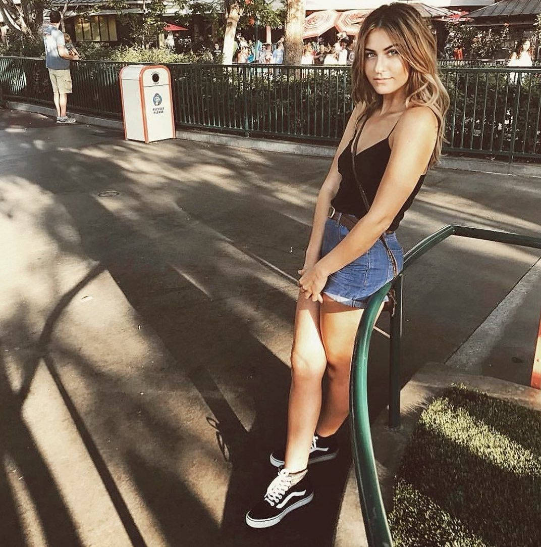 Scout Taylor-Compton 靠欄杆照 壁紙 — 一位身穿牛仔短褲和黑色上衣的年輕女性，在公園環境中，斑駁的陽光下，隨意地靠在綠色欄杆上。