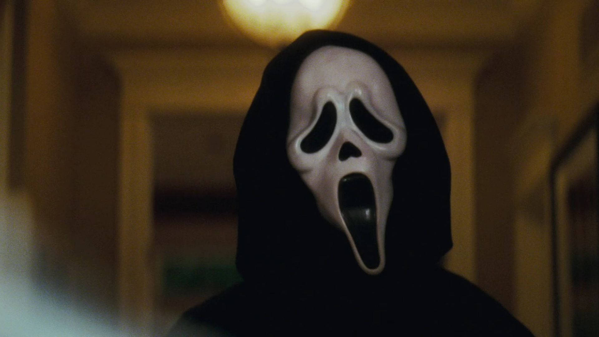 《Scream 4》中的殺手反派Ghostface 壁紙 — 一個戴著白色鬼面具（有黑色眼和嘴孔）的人影，站在昏暗的走廊裡，上方是溫暖的吊燈。