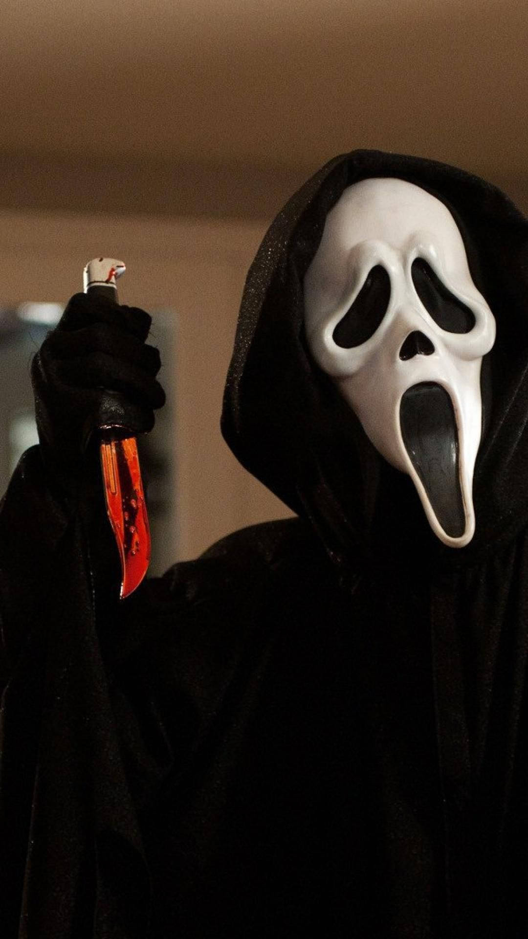 《Scream》中面具下的懸疑 壁紙 — 一個戴着白色Ghostface面具、身穿黑色連帽長袍的人，在燈光昏暗的房間裏拿着一把沾滿血的刀。