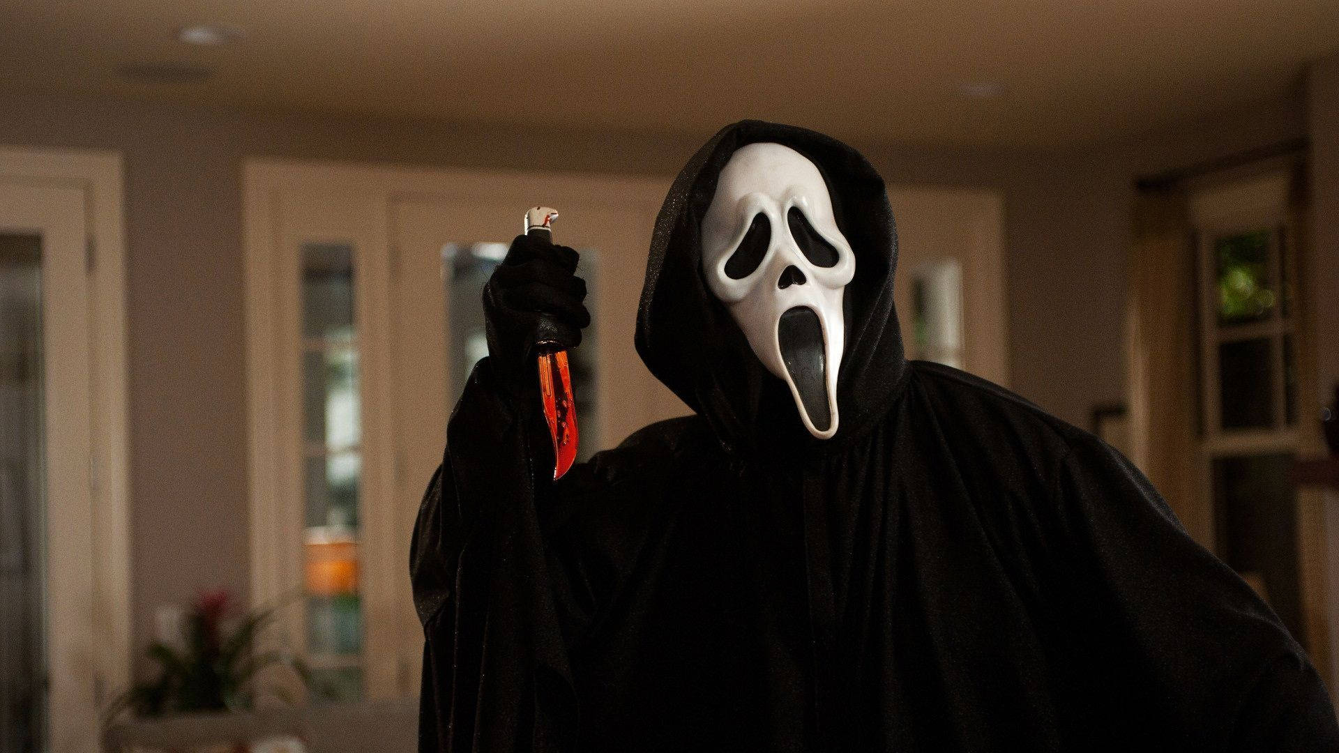 《Scream》電影場景 壁紙 — 一個身穿黑色連帽長袍、戴著標誌性白色鬼面具的人物，在昏暗的室內手持一把沾血的刀。