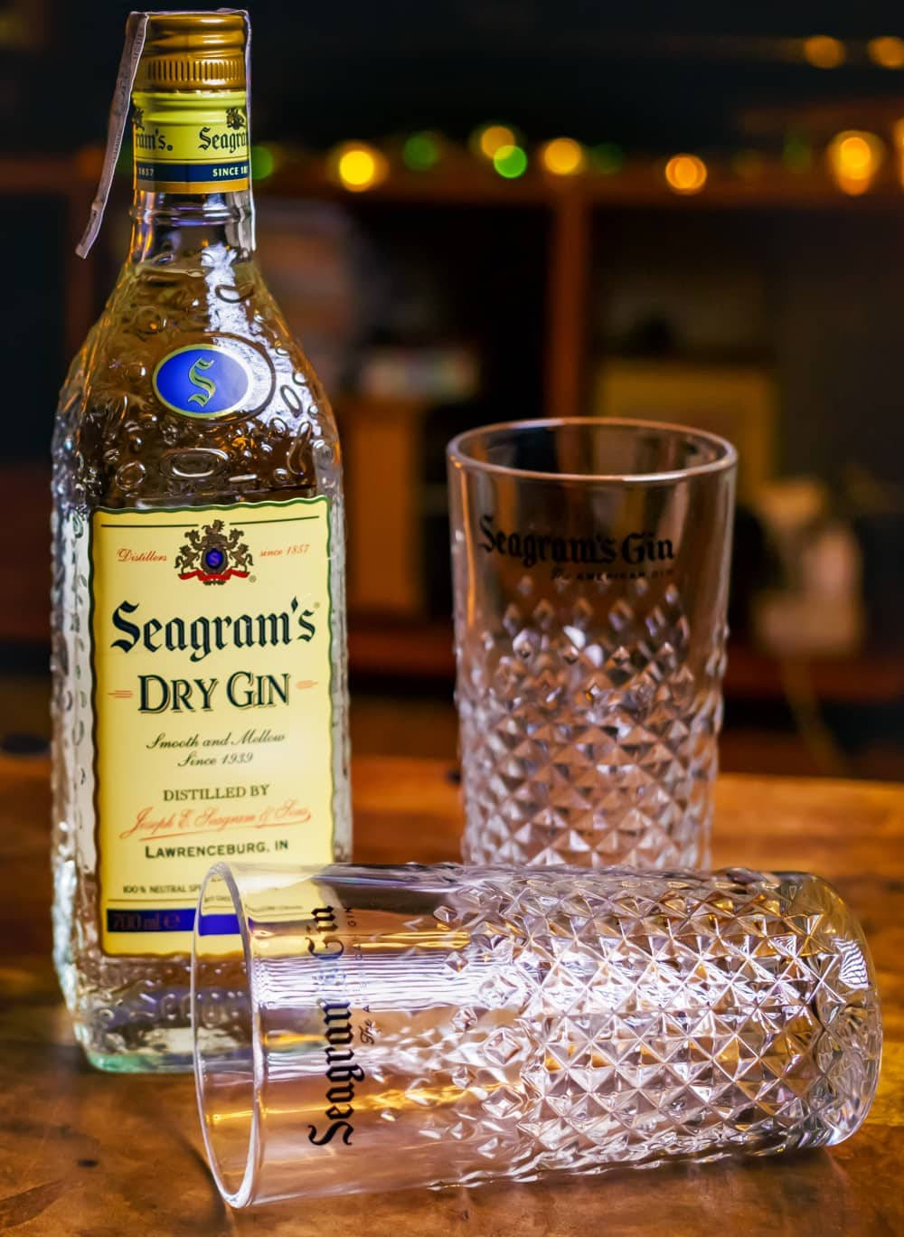 一瓶Seagram's 干金酒 壁紙 — 一瓶Seagram's Dry Gin和兩個帶圖案的玻璃杯放在木製吧檯上，背景有溫暖的散景燈光。