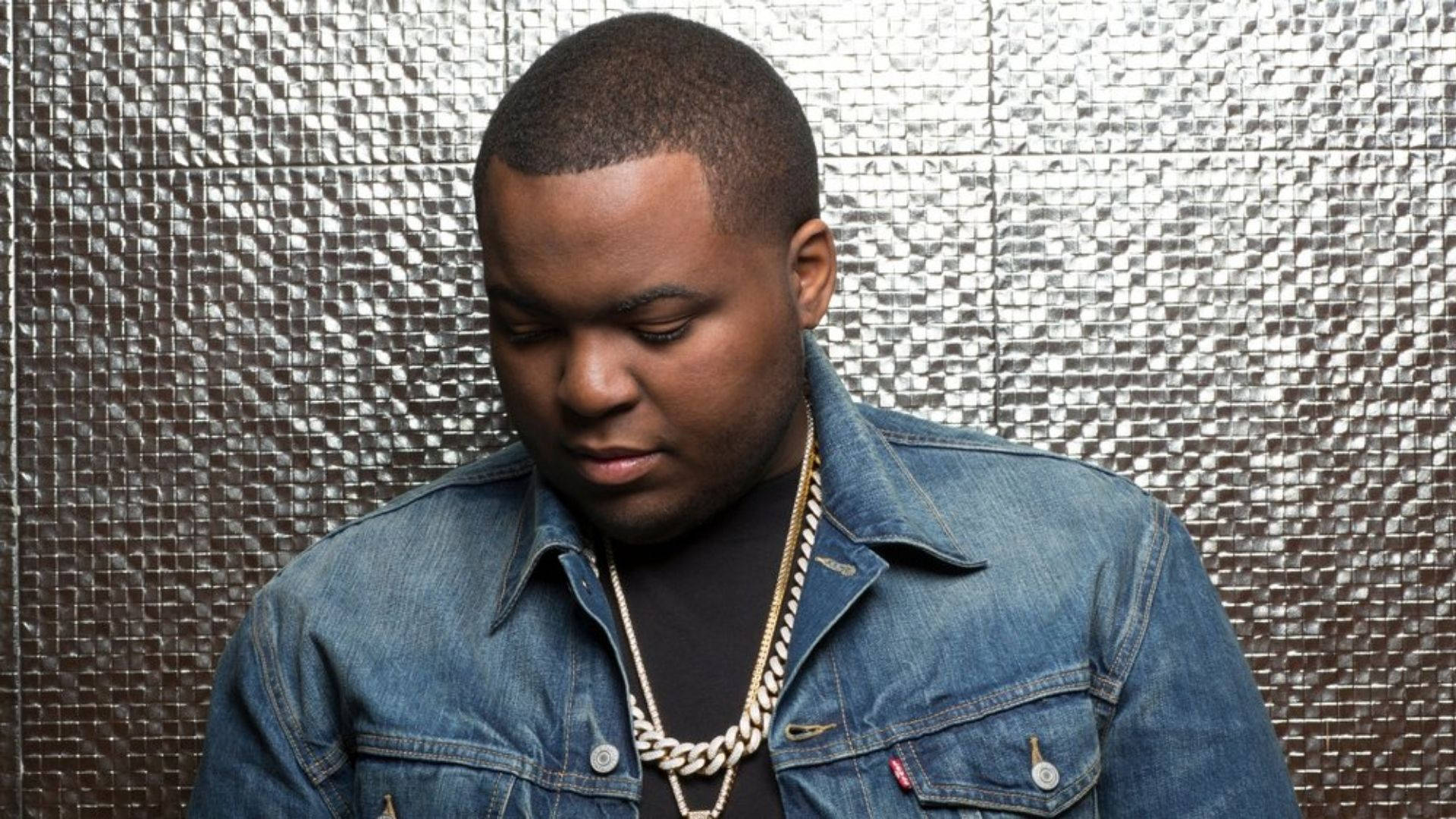 Sean Kingston 低頭酷姿 壁紙 — 一名身穿牛仔外套和金色鏈條的男子，在閃亮的金屬牆前低頭。