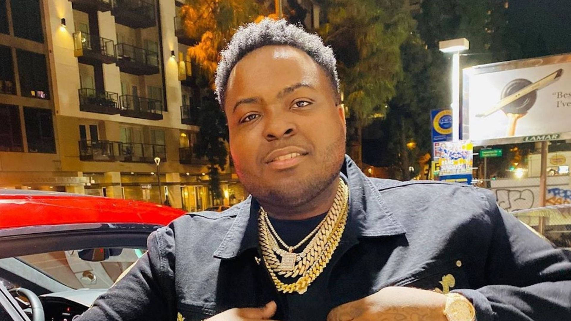 Sean Kingston 樂趣夜晚 壁紙 — 一個擁有時尚髮型和金鏈的男人在城市的夜燈下自信地擺姿勢，旁邊是一輛紅色汽車。