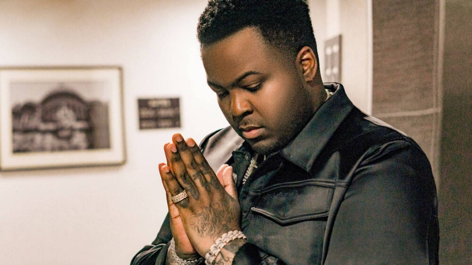 Sean Kingston 美學姿勢 壁紙 — 一名身穿黑色外套的男子在光線柔和的室內房間裡雙手合十祈禱，牆上掛有裱框藝術品。