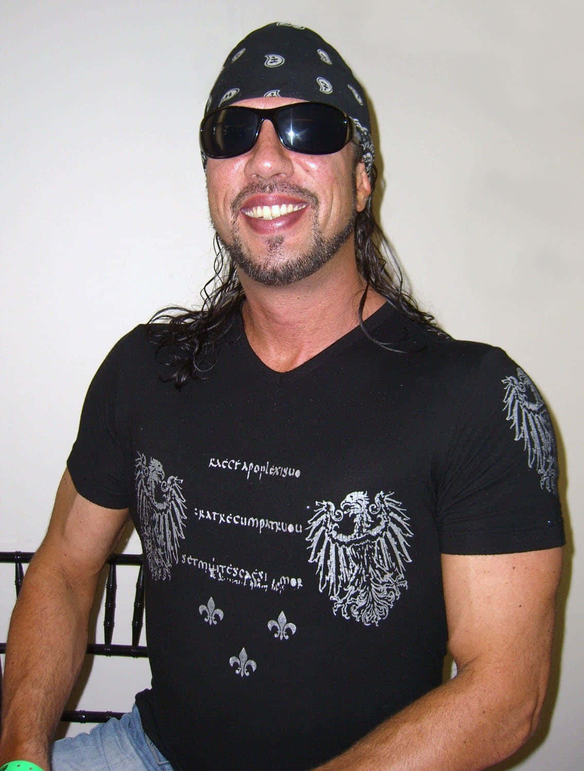 戴太陽眼鏡的摔角手Sean Waltman 壁紙 — 一位微笑的長髮男子戴著太陽眼鏡，穿著印有老鷹和百合花圖案的黑色T恤，坐在室內。