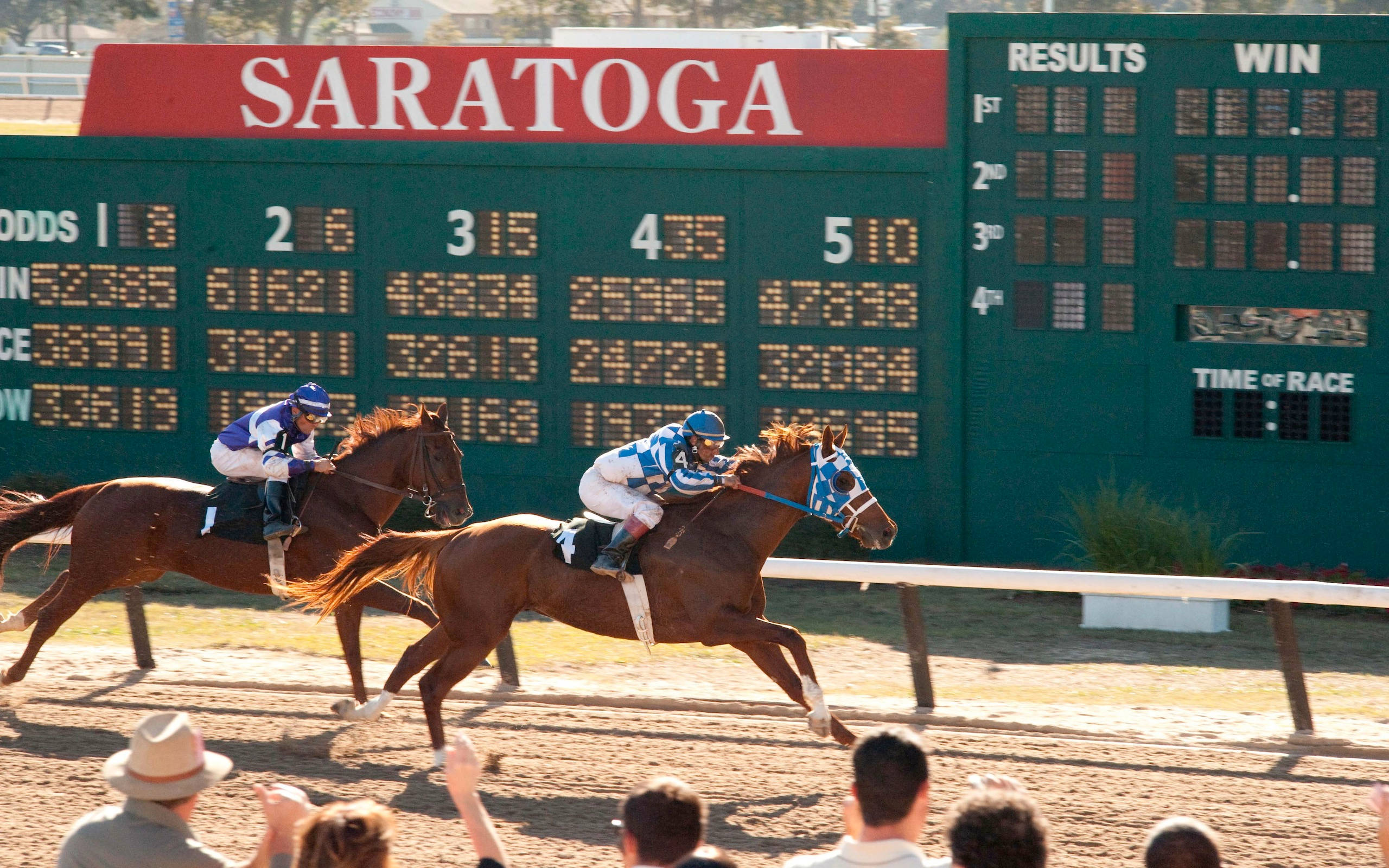 Secretariat賽馬場Saratoga 壁紙 — 騎師在泥地賽道上策騎棕色馬匹，身後有綠色的薩拉托加計分板顯示賠率和結果。