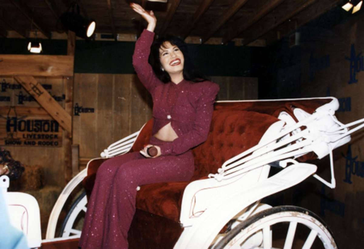Selena Quintanilla 騎馬車 壁紙 — 一位身穿閃亮栗色連身褲的女性在牲畜展上從白色馬車上愉快地揮手。