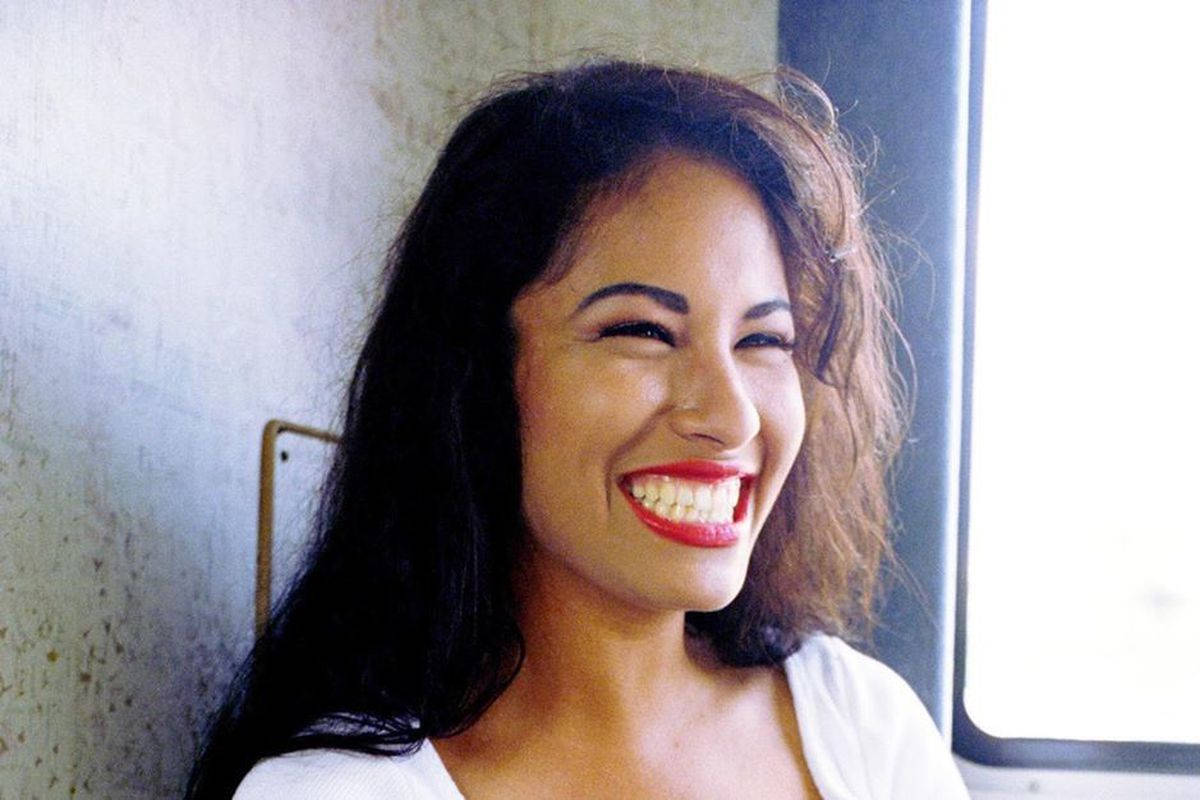 Selena Quintanilla 無敵笑容 壁紙 — 一位深色頭髮、塗著鮮紅色唇膏的女性，靠在窗邊有紋理的牆上，開懷大笑。