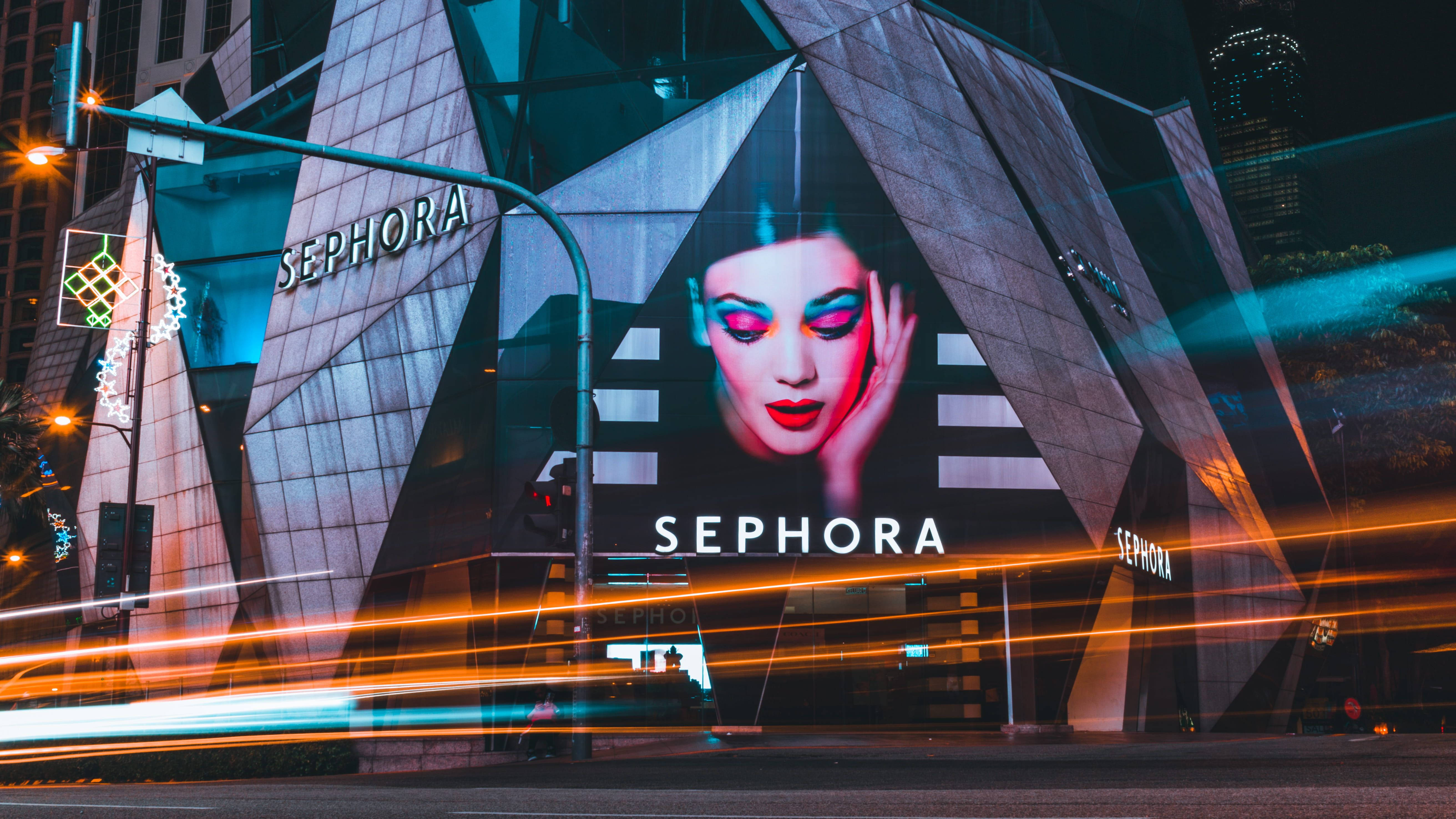 Sephora 美妝零售店 壁紙 — 夜晚發光的Sephora廣告牌展示了一位女性臉龐，妝容大膽，背景是幾何建築外牆和流動的城市燈光。