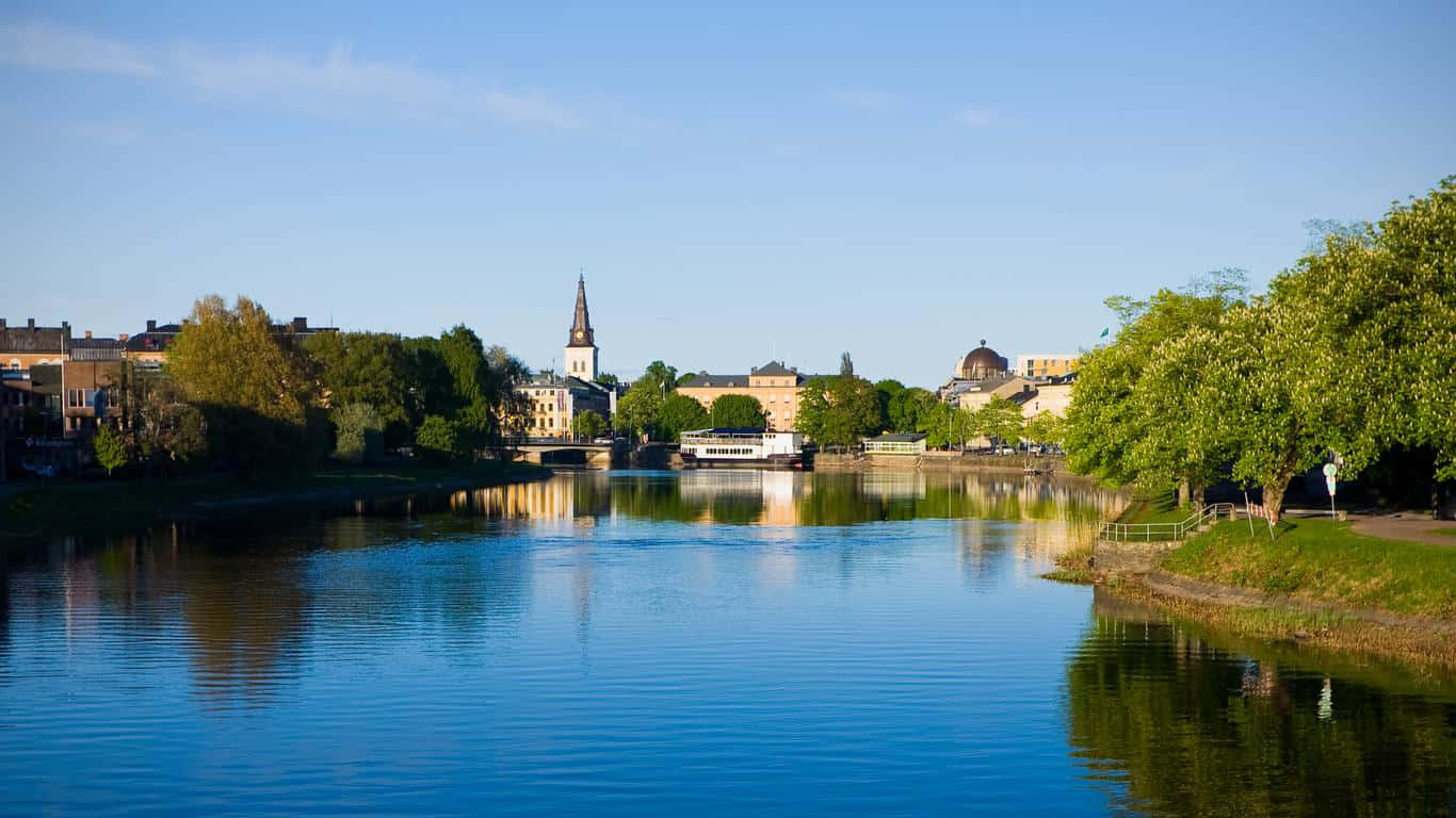 Karlstad Sweden 桌布 壁紙 — 一條寧靜的河流映照著歷史建築和茂密的綠樹，在清澈的藍天下。