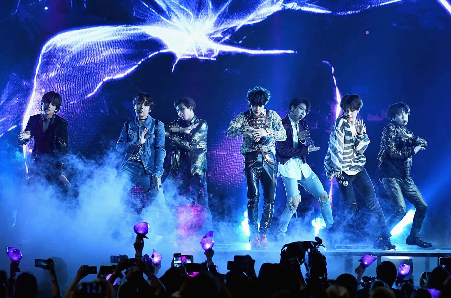 K-pop 組合在燈光璀璨的舞台上表演，現場有煙霧和手持螢光棒的粉絲。