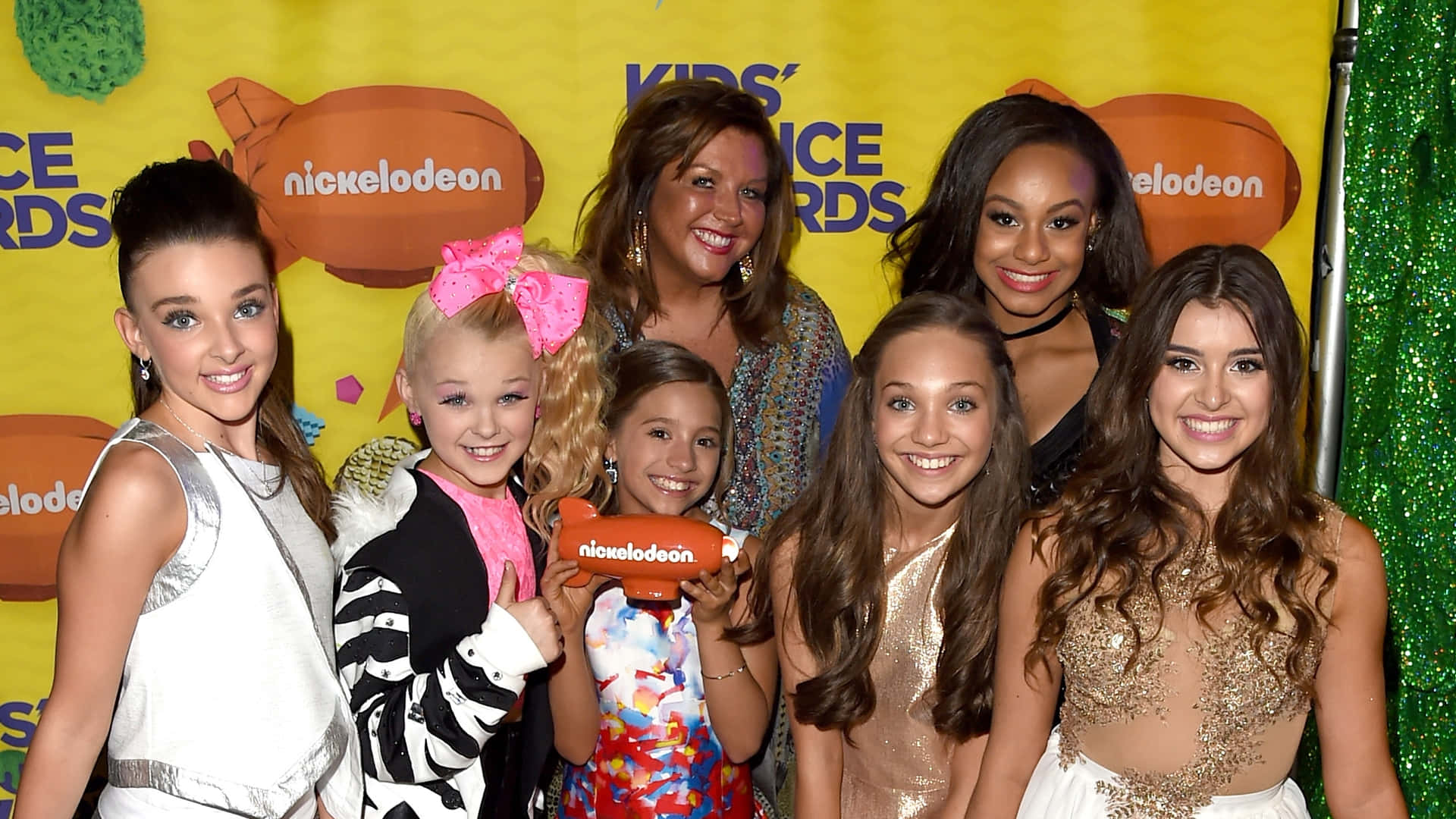  壁紙 — 七個微笑的年輕女孩和一個女人在Nickelodeon Kids' Choice Awards上一起擺姿勢，背景是明亮的黃色和綠色閃粉牆。