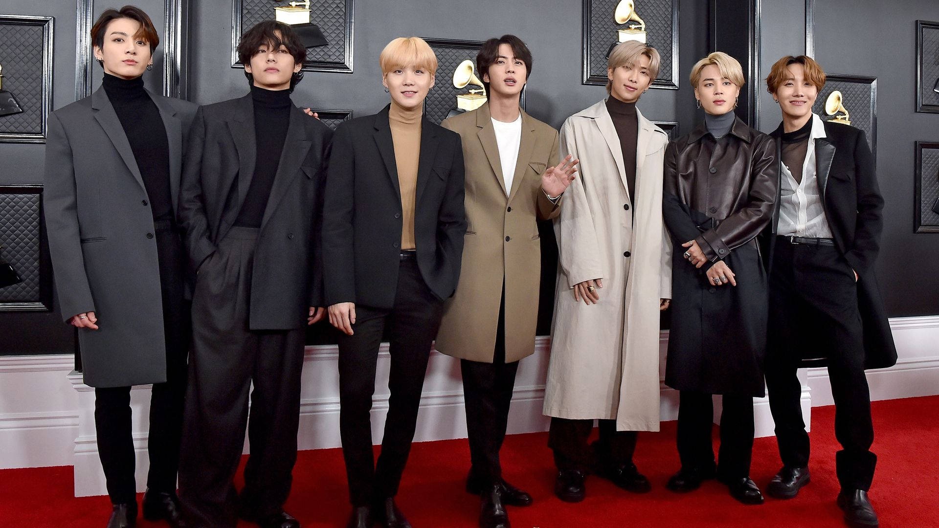 BTS 紅毯桌面壁紙 壁紙 — 七位穿著訂製大衣的時尚男士在黑色牆壁前、金色格林美獎盃襯托下的紅地毯上自信地擺姿勢。