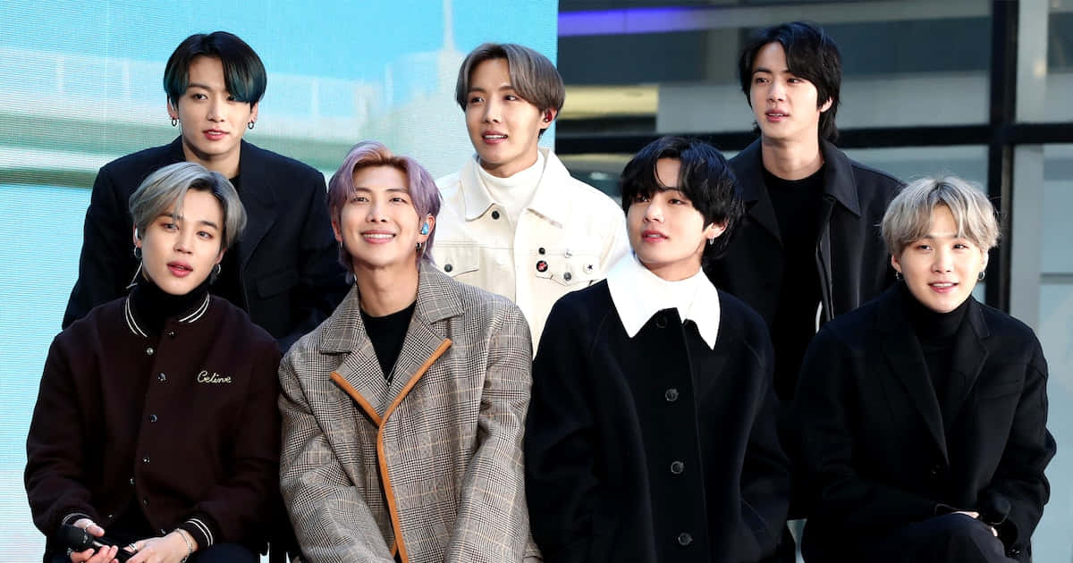 BTS 在NBC Today Show訪問照 壁紙 — 七位時尚年輕男士穿著潮流外套，在現代玻璃建築背景下自信地擺姿勢。