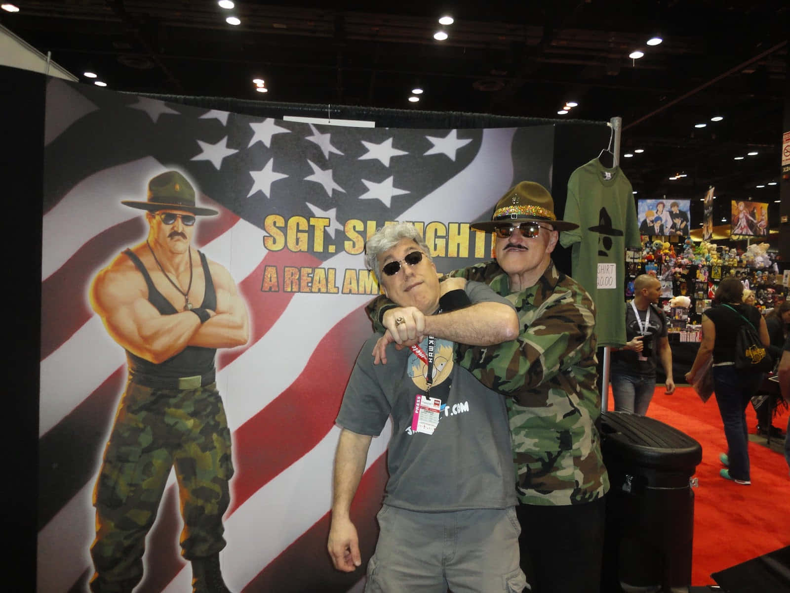 Sgt Slaughter 在Chicagoland漫畫與娛樂展展示招牌動作 壁紙 — 兩名戴迷彩帽和太陽眼鏡的男子在Sgt. Slaughter漫畫展背景前自信地擺姿勢。