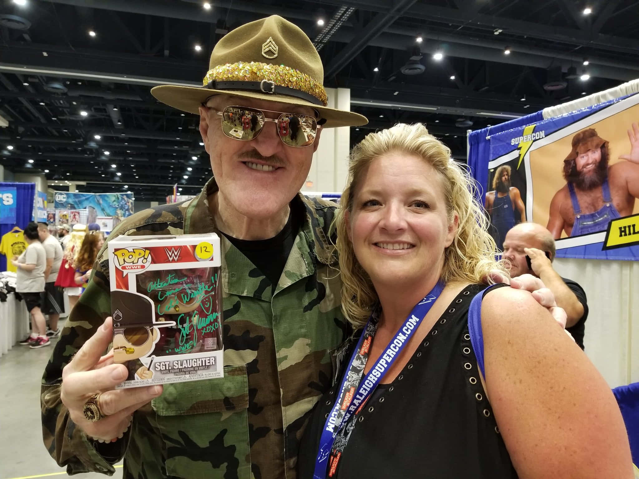 Sgt Slaughter 在SuperCon與粉絲合影 壁紙 — 一個戴著軍帽和太陽眼鏡的微笑男人，拿著一個簽名的WWE Funko Pop，與一個女人在會議上擺姿勢。