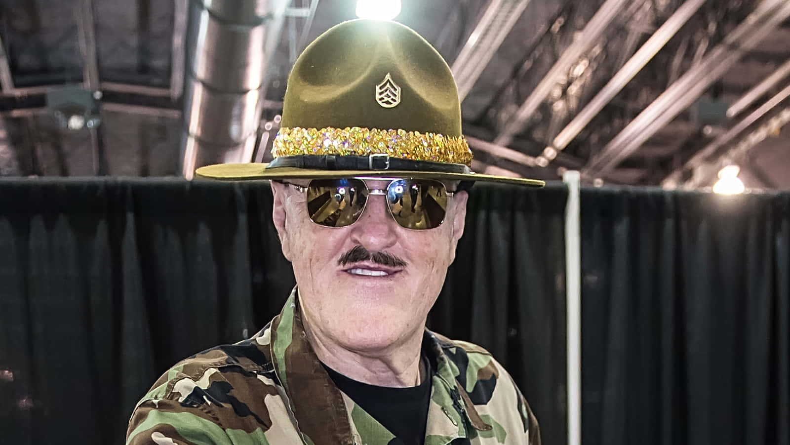 Sgt Slaughter 摔角先驅 壁紙 — 一名身穿迷彩服和裝飾軍帽的男子在室內戴著反光太陽眼鏡微笑。