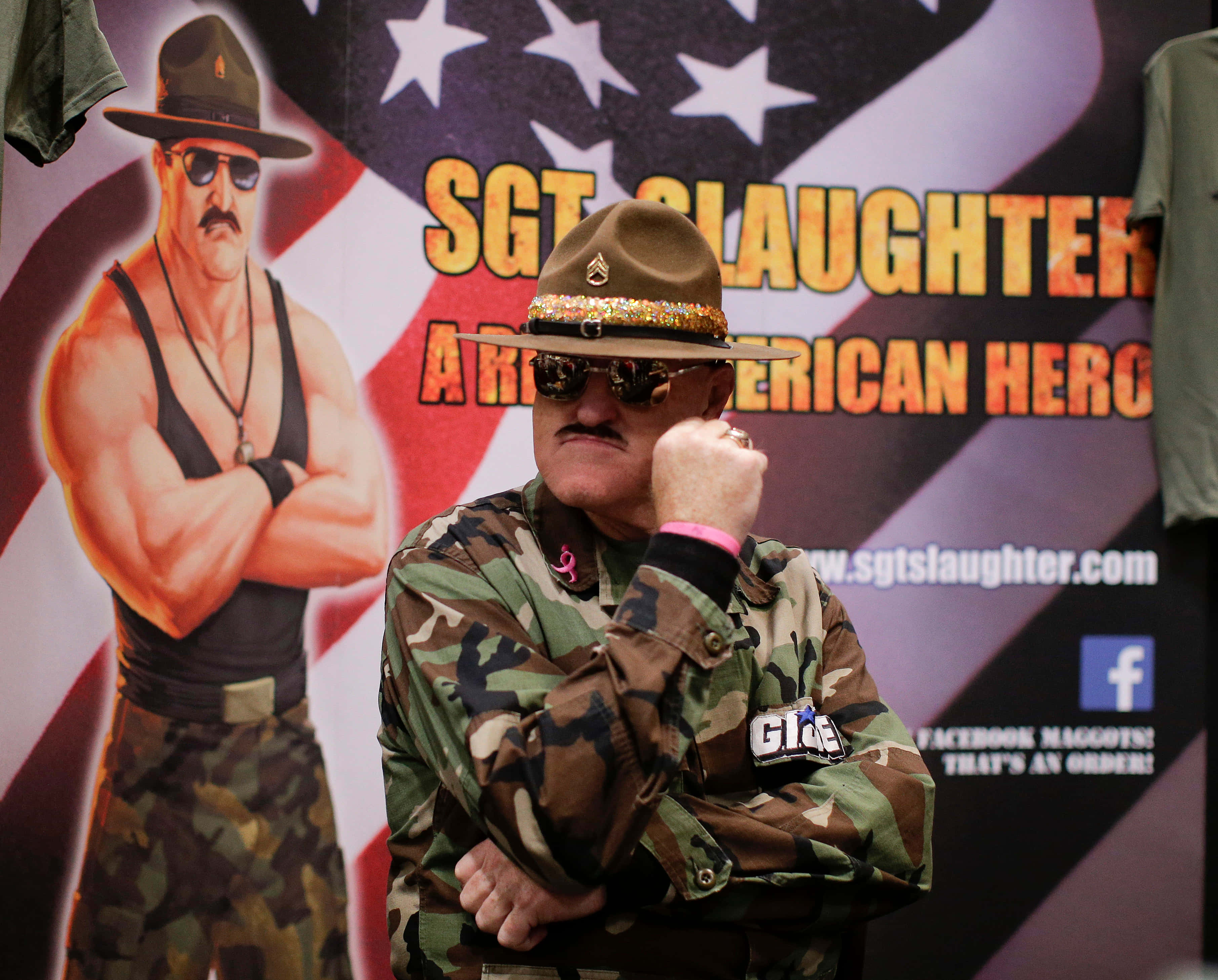 Sgt Slaughter 摔角傳奇 壁紙 — 一名身穿迷彩服和裝飾帽子的男子自信地在帶有美國國旗圖案的Sgt Slaughter摔角海報前擺姿勢。