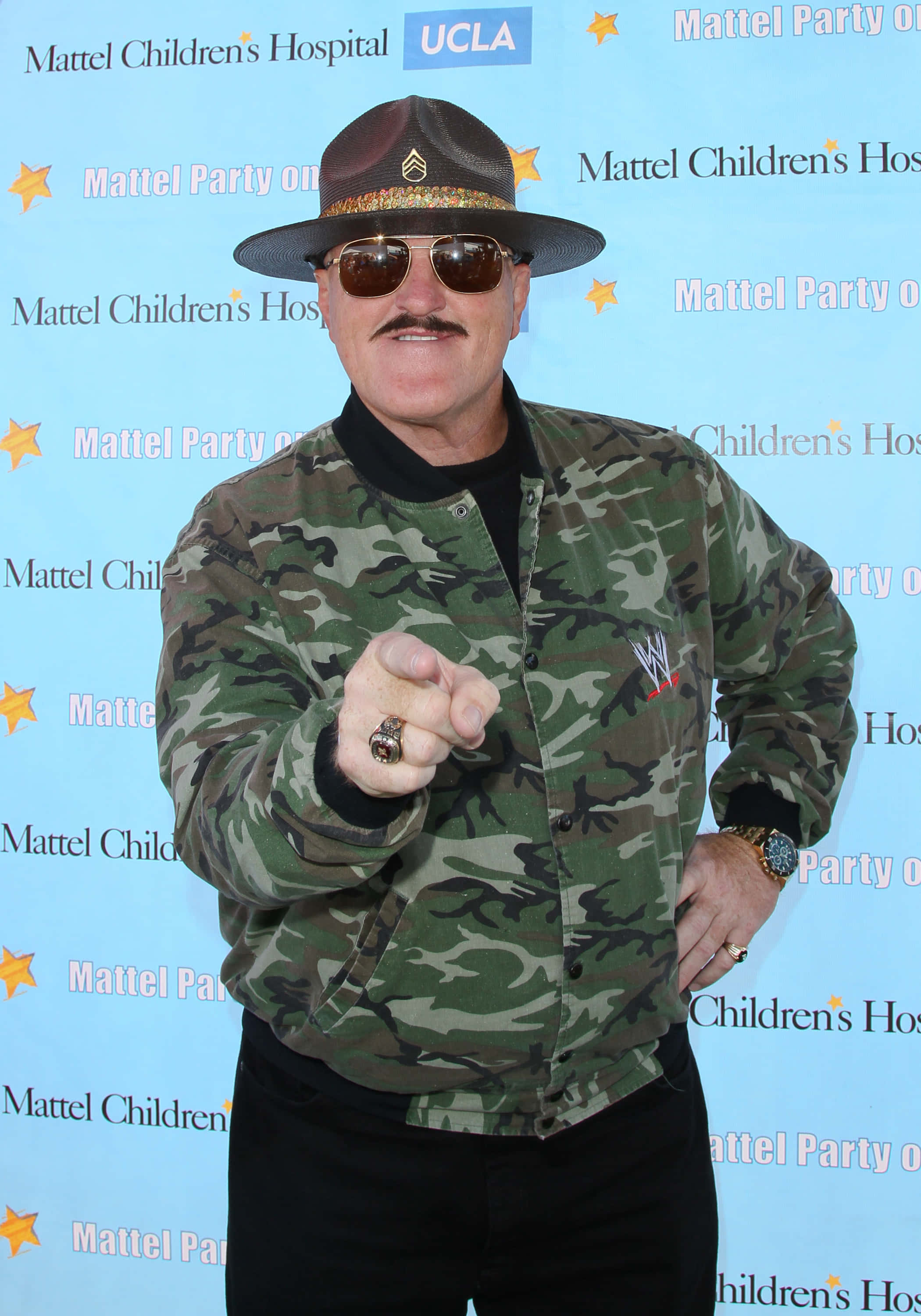 Sgt Slaughter 第12屆Mattel派對 壁紙 — 一名身穿迷彩外套和軟呢帽的男子，在藍色活動背景前自信地指向相機。