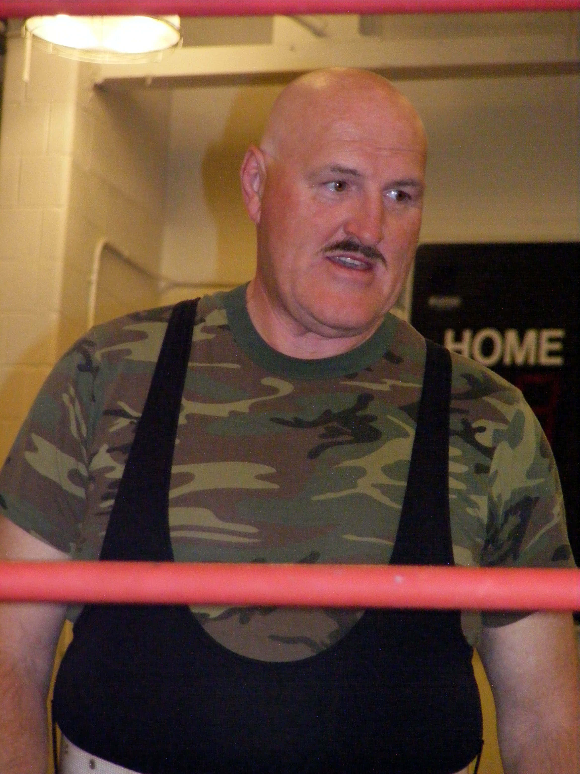 Sgt Slaughter 退役摔角巨星 壁紙 — 一位光頭留鬍鬚的摔角手穿著迷彩襯衫，站在紅色邊框的摔角擂台內，在明亮的燈光下。