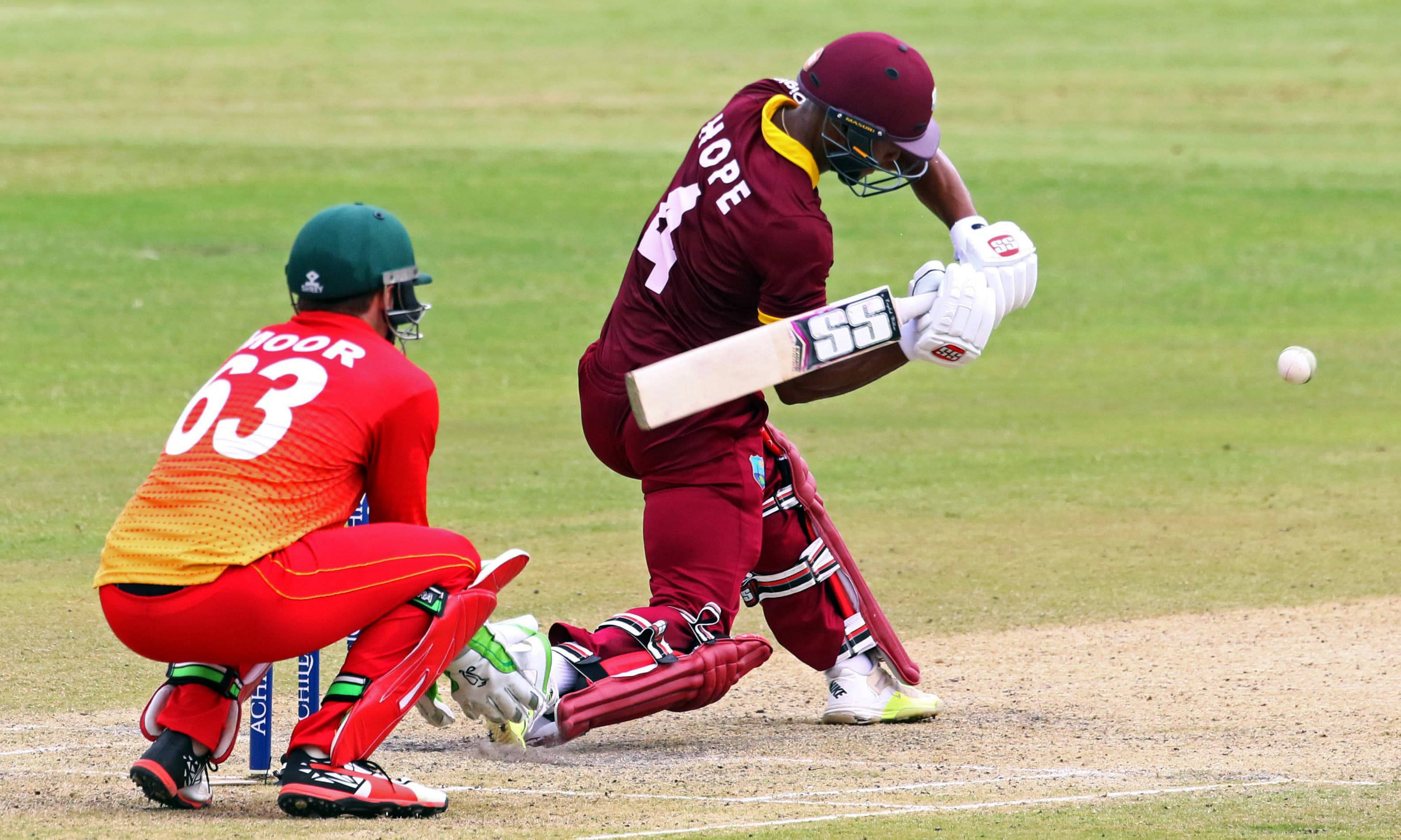 Shai Hope 擊球姿勢 壁紙 — 一名身穿栗色球衣的板球擊球手揮棒擊球，而一名身穿紅色球衣的守門員蹲在他身後的綠色場地上。