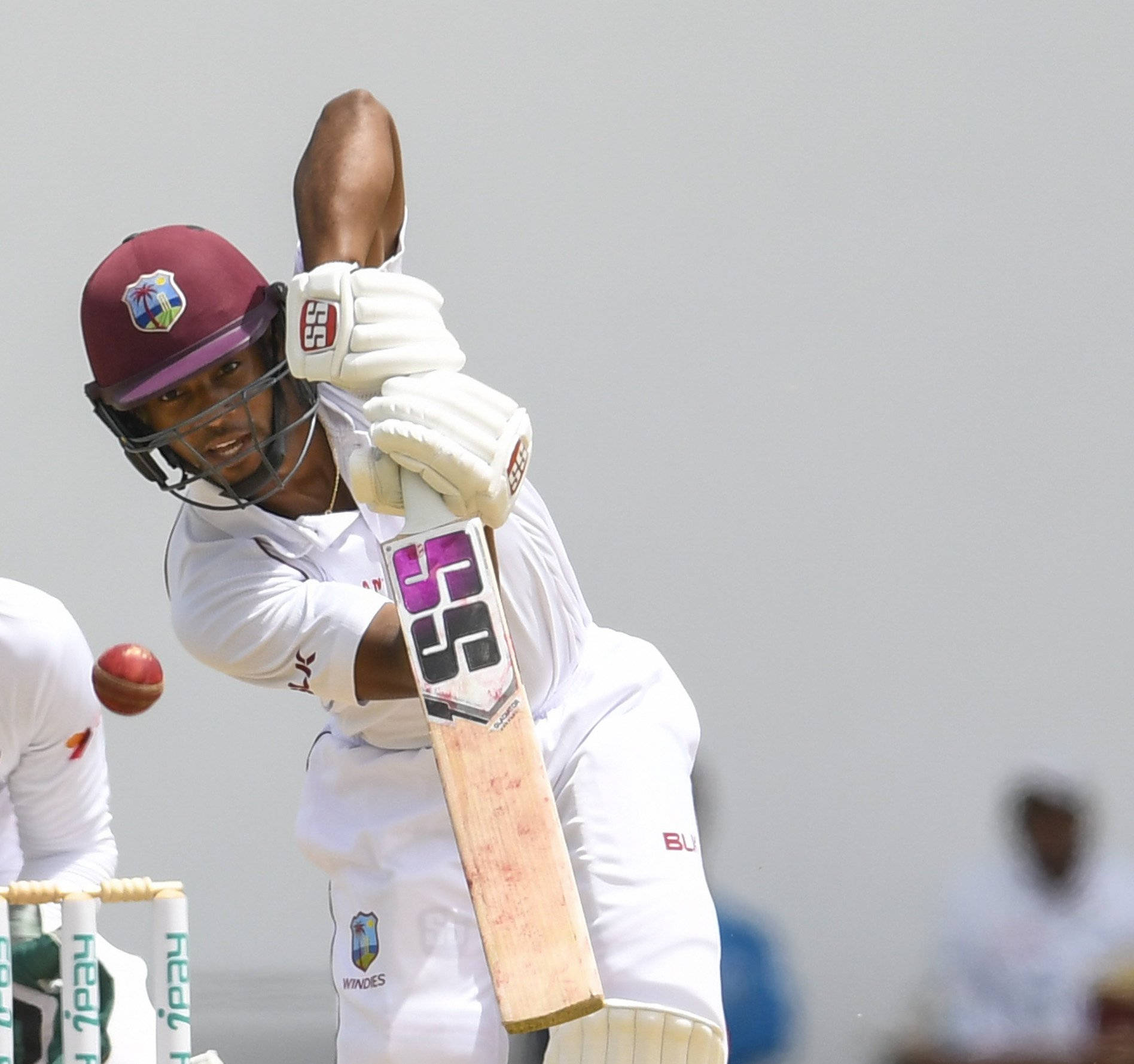 Shai Hope 扭轉握棒姿勢 壁紙 — 一名身穿白色制服和栗色頭盔的板球擊球手在日間比賽中揮動球棒擊打紅球。
