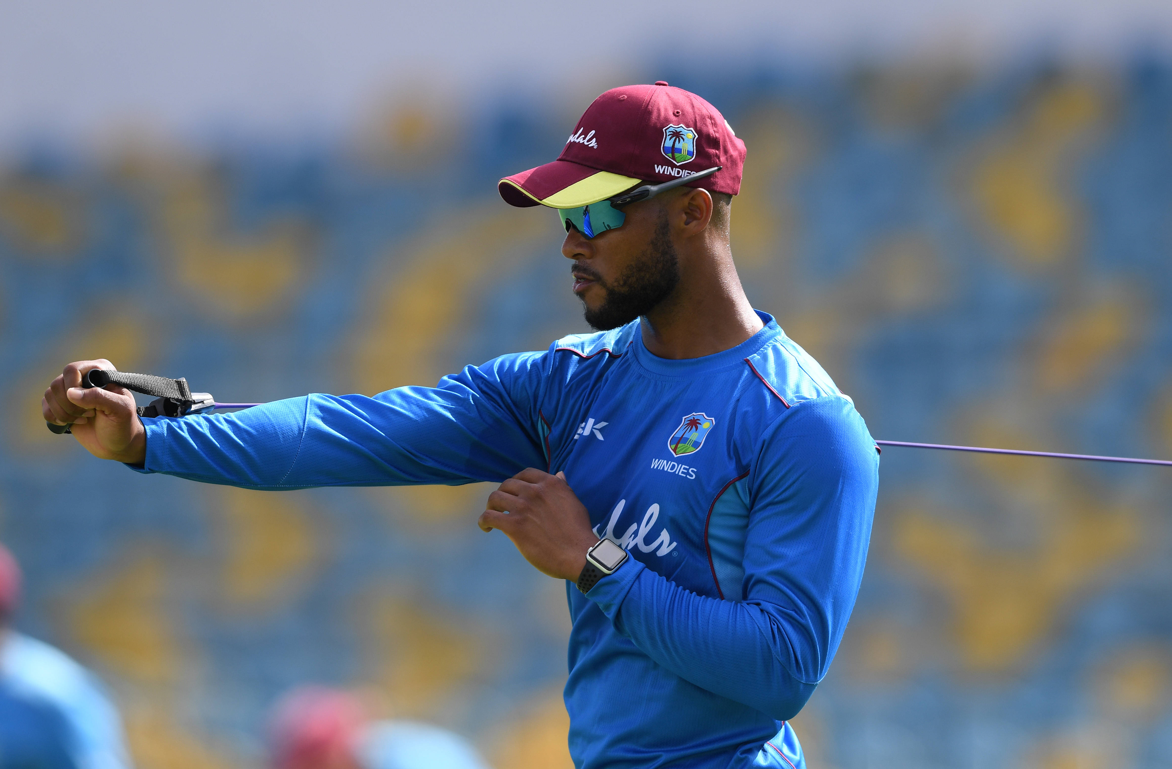 Shai Hope 拉伸手臂 壁紙 — 一位西印度群島板球運動員穿著藍色訓練服，在體育場明亮的日光下用阻力帶伸展。