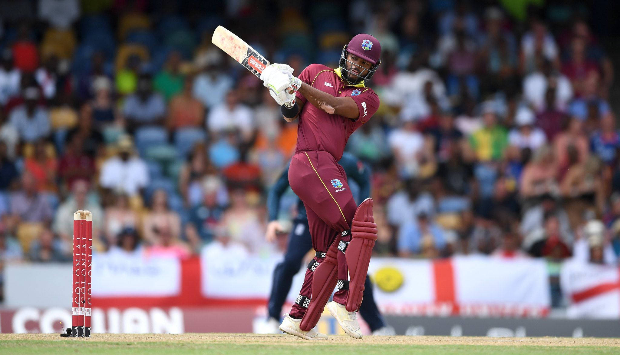 Shai Hope 擊球動作 壁紙 — 一名身穿棗紅色球衣的板球運動員在日間比賽中，在人群背景下強力揮棒擊球。