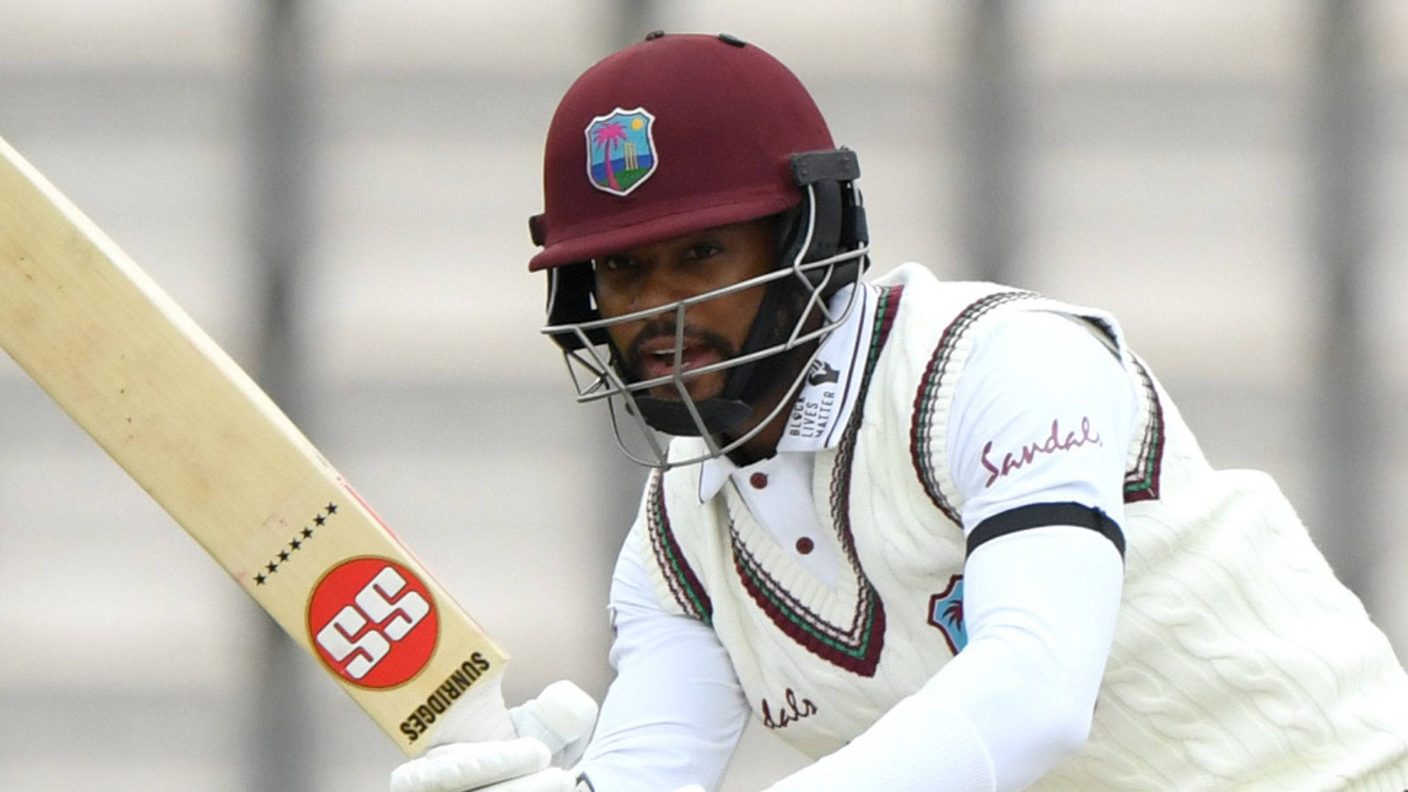 Shai Hope 板球造型 壁紙 — 一名身穿栗色頭盔和白色制服的板球擊球手，在揮棒中途緊握球棒，神情專注。