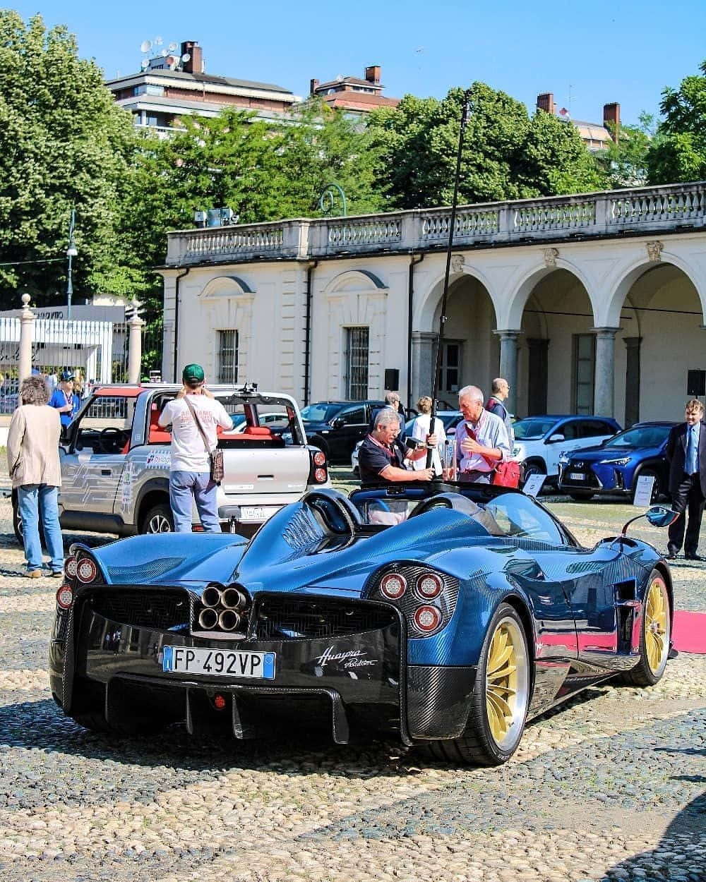 Pagani Zonda C12 Roadster 桌布 壁紙 — 一輛閃亮的藍色帕加尼Huayra跑車配金色輪胎停在鵝卵石路上，人們在古典建築附近欣賞它。